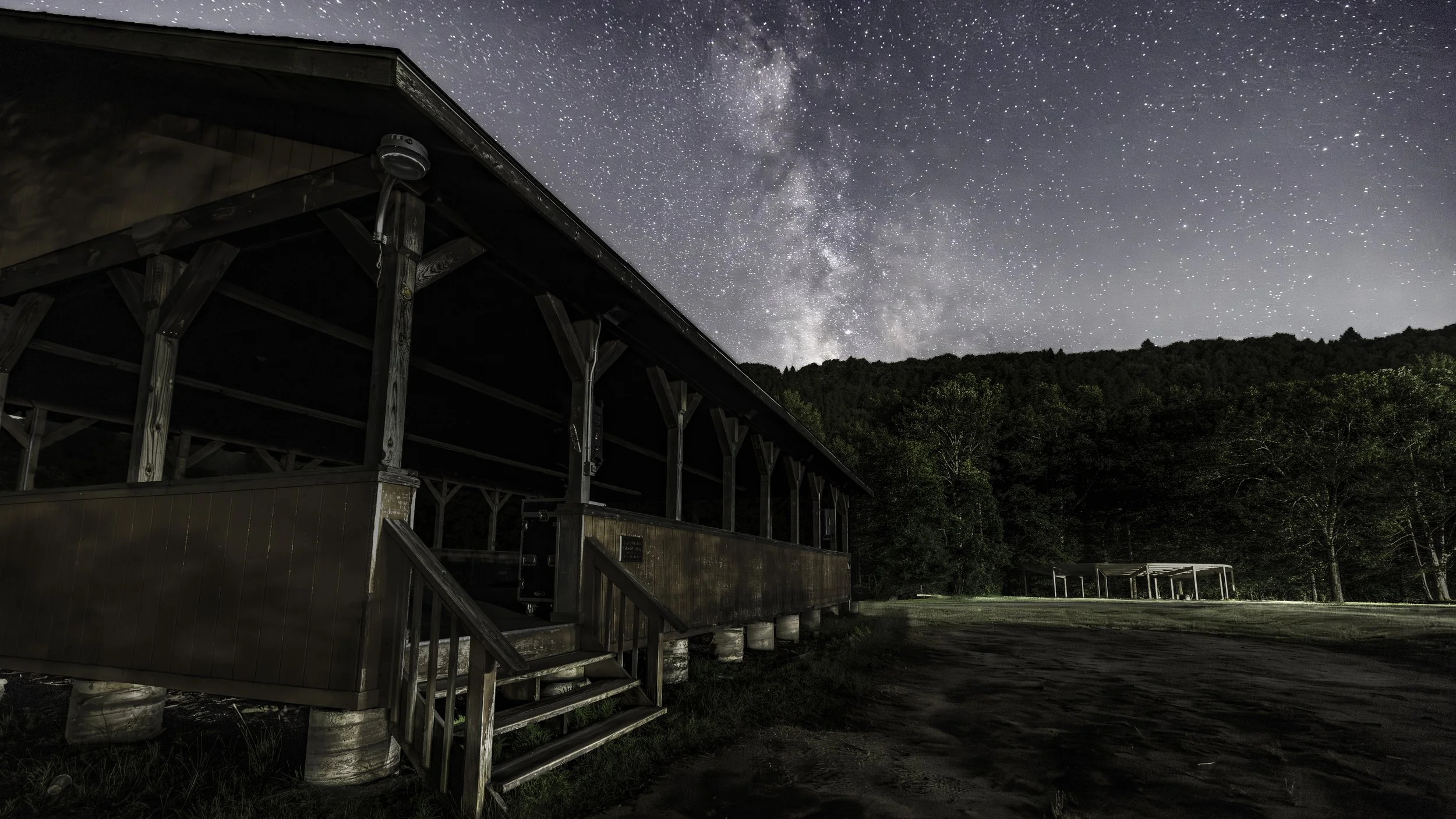 A wooden pavilion at night under a starry sky, with the Milky Way galaxy visible, surrounded by trees and an open field.