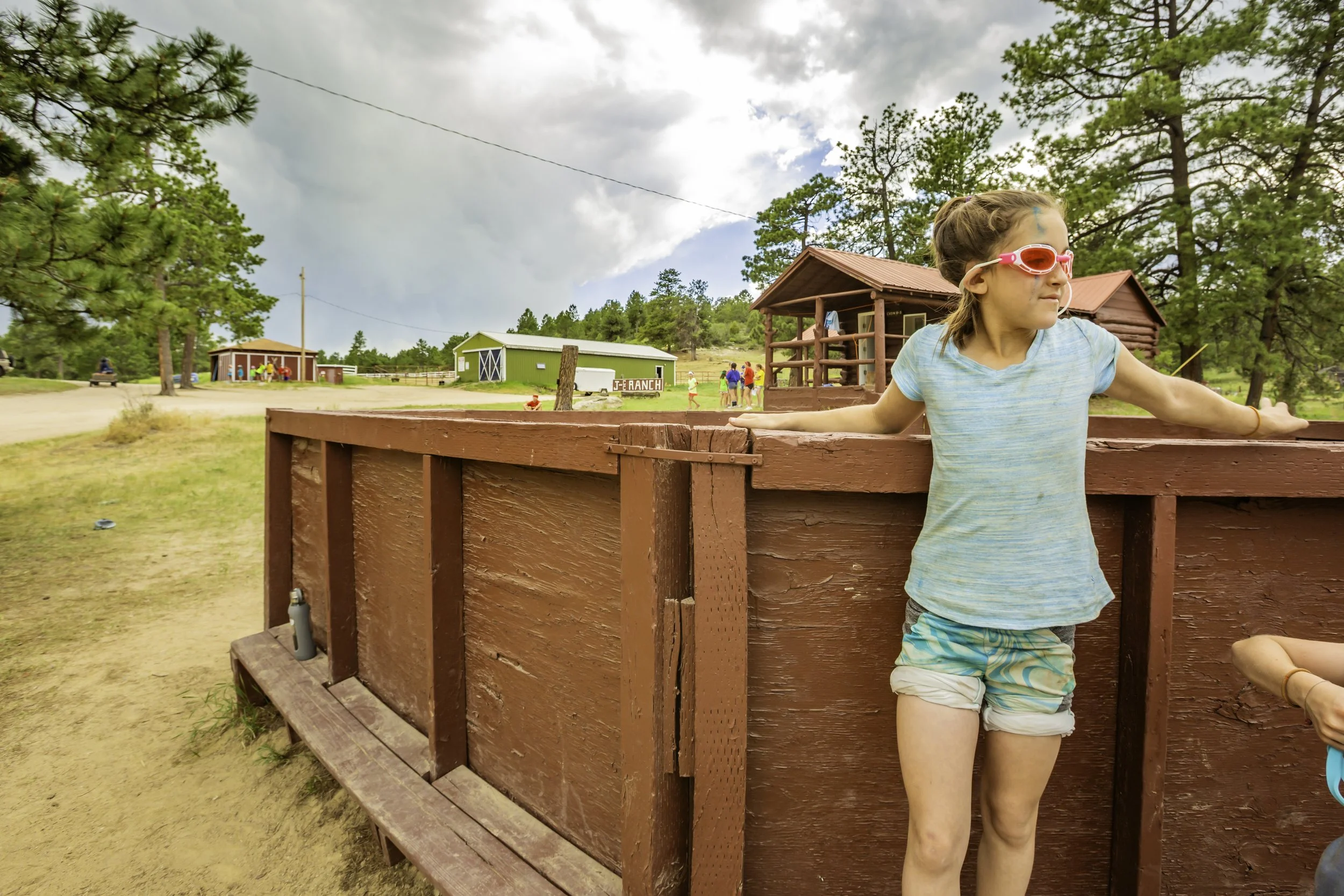 A girl with sunglasses and a striped shirt leaning on a wooden corral at a ranch, with barns and kids in the background under cloudy skies.