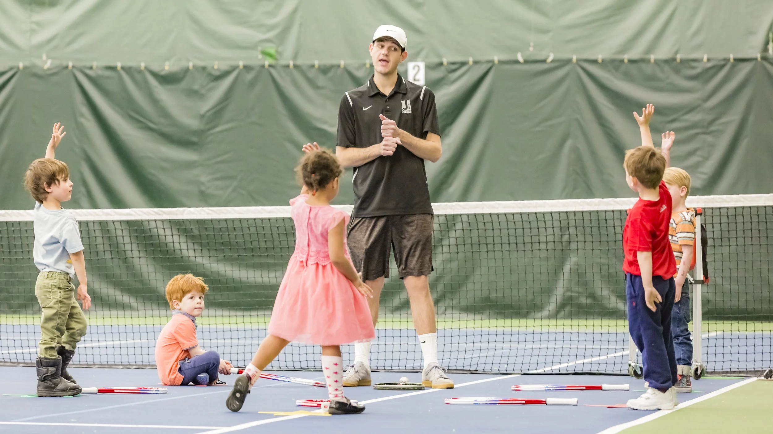 A young male tennis coach standing on an indoor tennis court, giving instructions to a group of five children, some of whom are raising their hands.