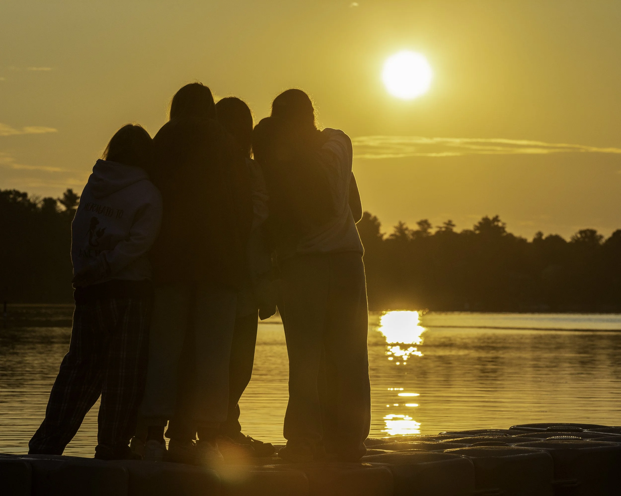 People standing on a dock at sunset over a body of water, with trees in the distance.