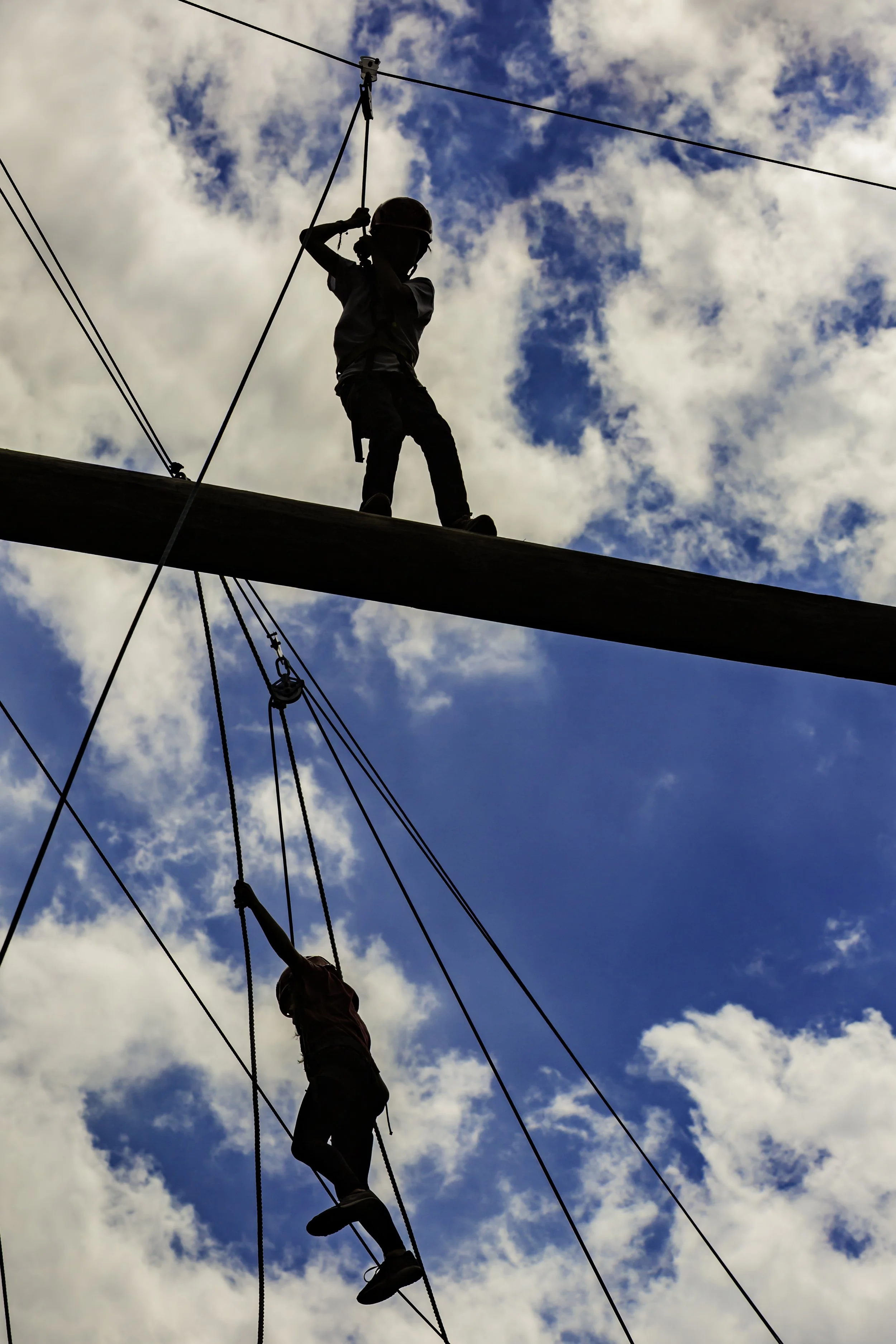 Two children climbing a high ropes course against a partly cloudy sky.