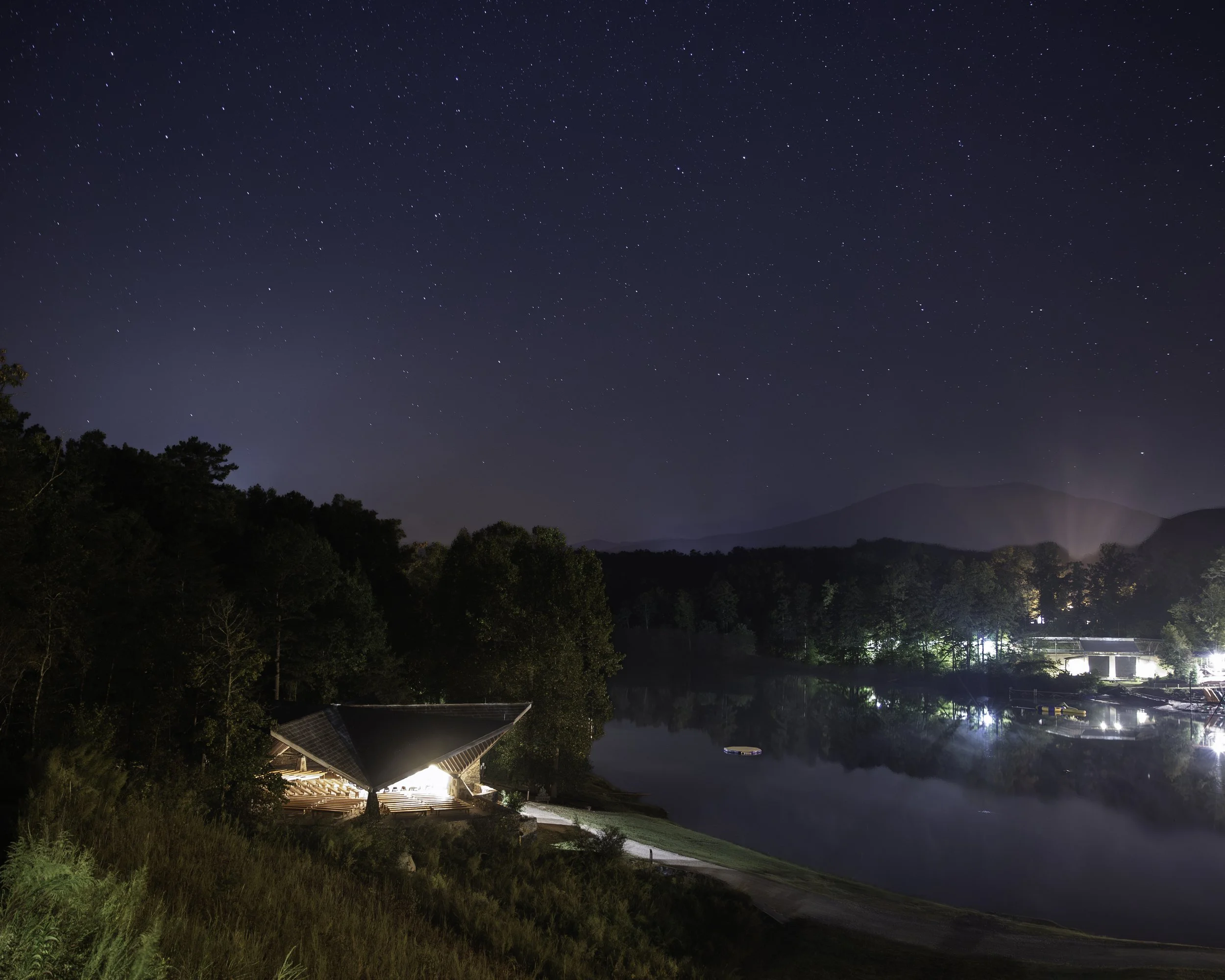 Nighttime scene of a lake with trees on the shoreline, reflecting lights from buildings, under a starry sky with mountain silhouettes in the background.