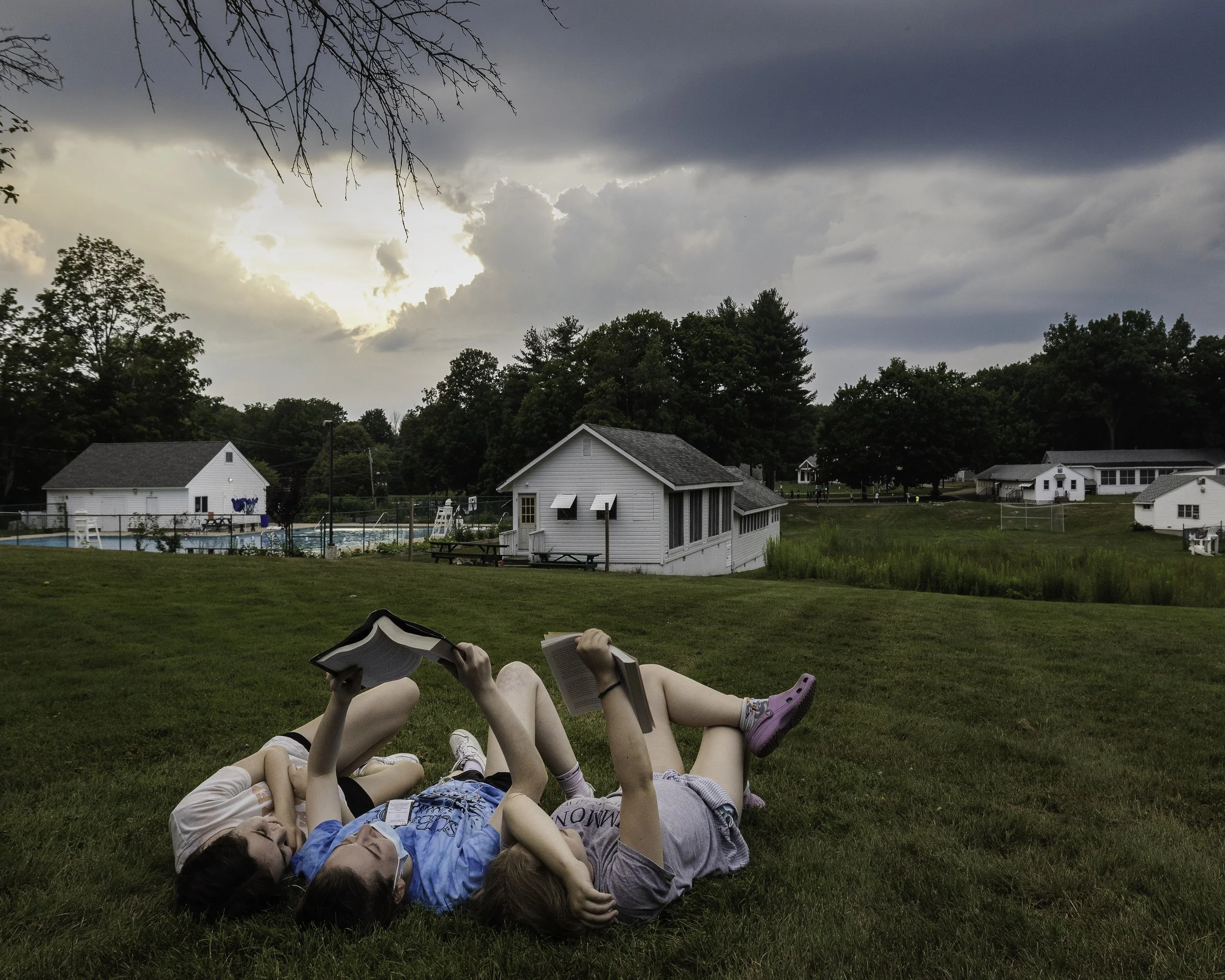 Three kids lying on grass, reading books, with a pool, houses, and trees in the background during cloudy weather.
