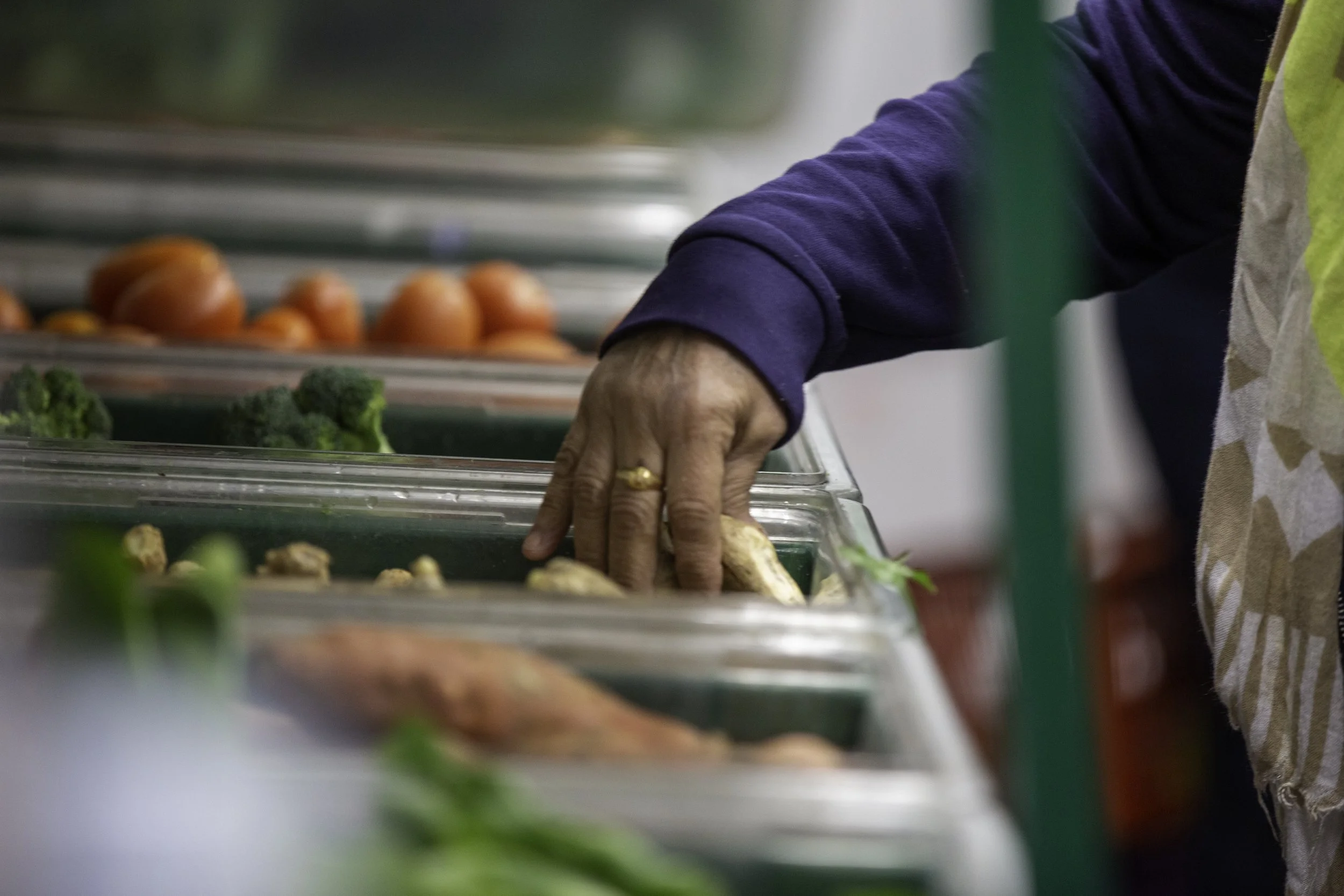 Close-up of a person's hand reaching into a glass food display case filled with vegetables like broccoli, tomatoes, and ginger at a market.