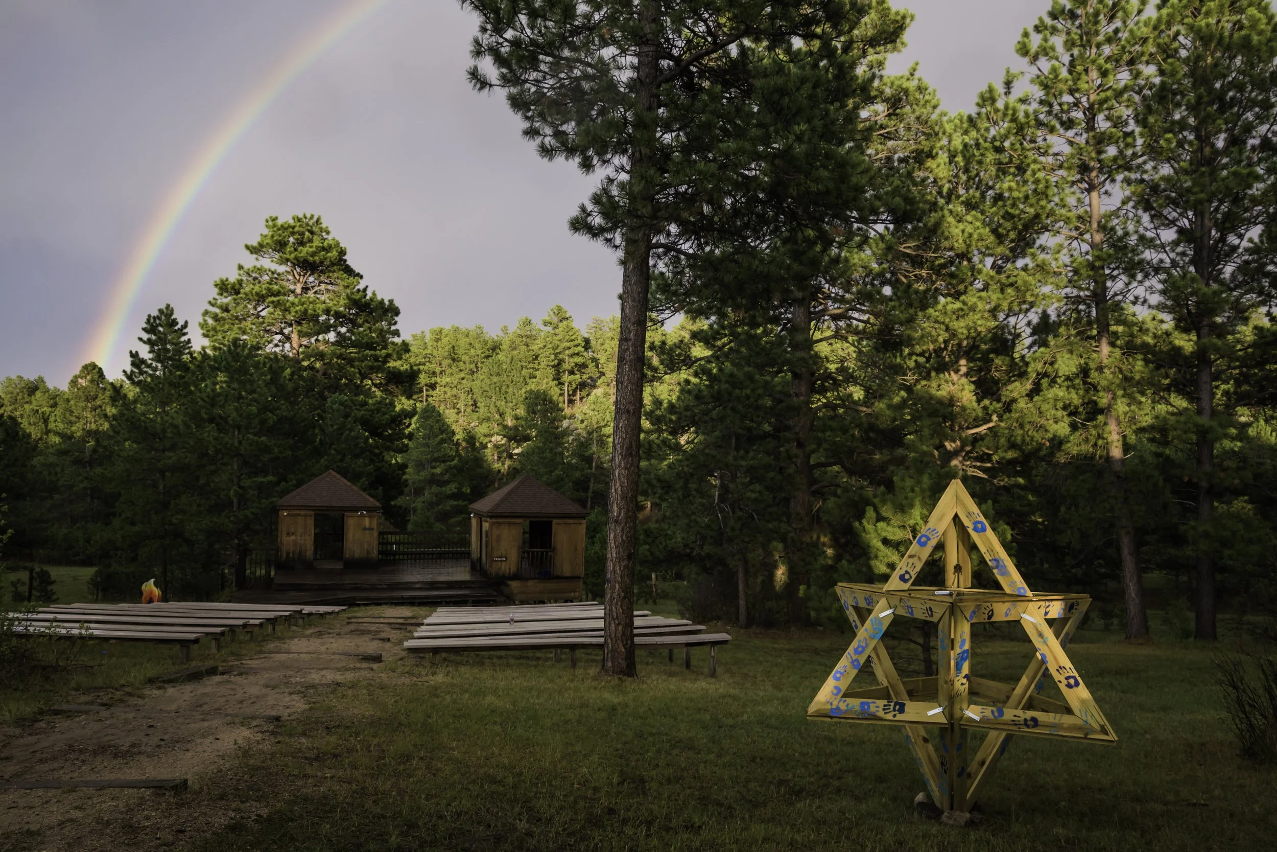 A rainbow arches across a cloudy sky over a wooded outdoor area with tall pine trees. In the foreground, there's a wooden star-shaped puzzle and a grassy clearing with some wooden platforms. Two small wooden structures with pitched roofs are visible 