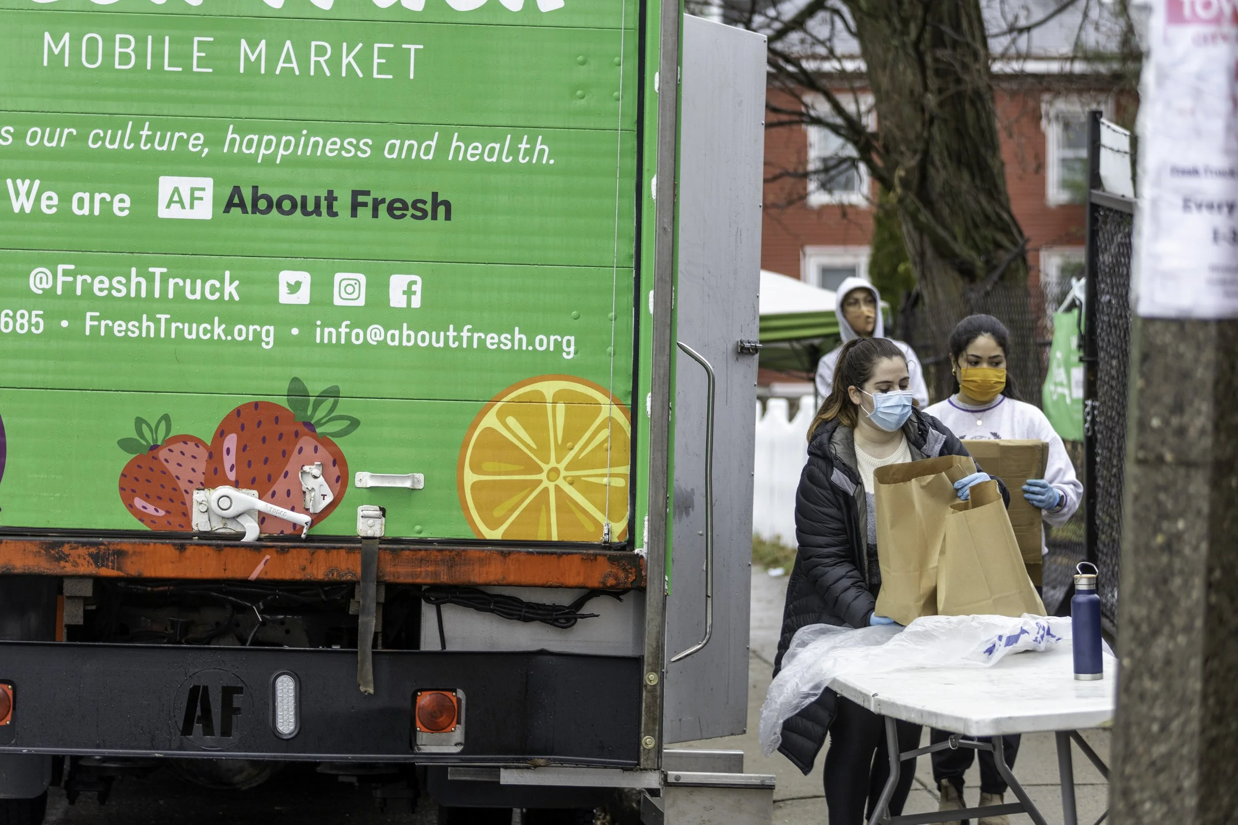 People wearing masks placing groceries in paper bags at an outdoor mobile market. A green truck with fruit illustrations and the words 'About Fresh' and contact information is visible.