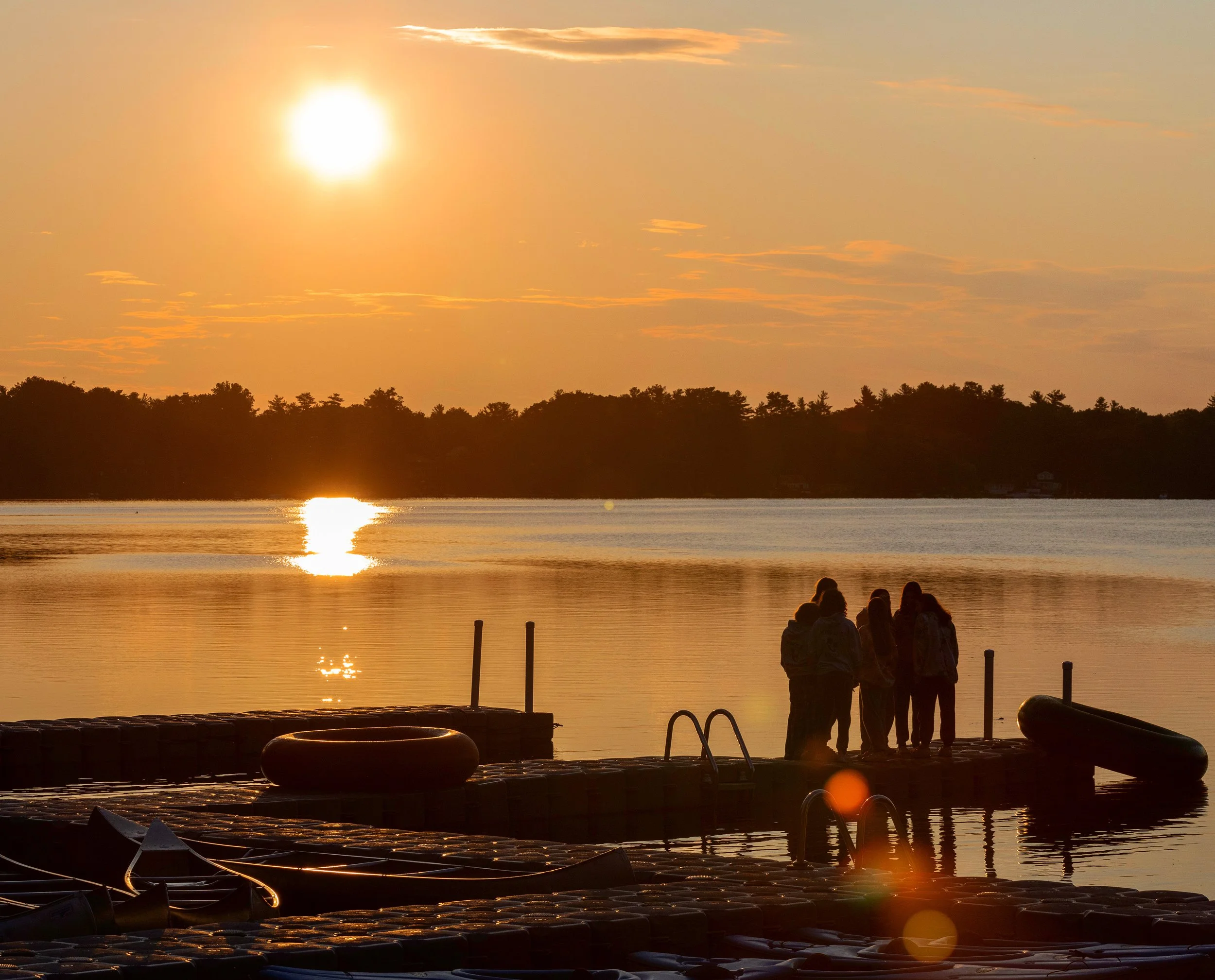 A group of people standing on a dock by a calm lake during sunset, with boats docked nearby and the sun reflecting on the water.