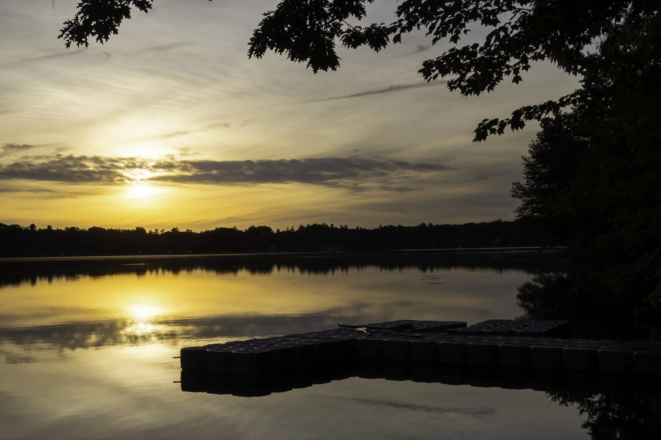 Sunset over a calm lake with reflections, trees and clouds in the sky, and a dock in the foreground.