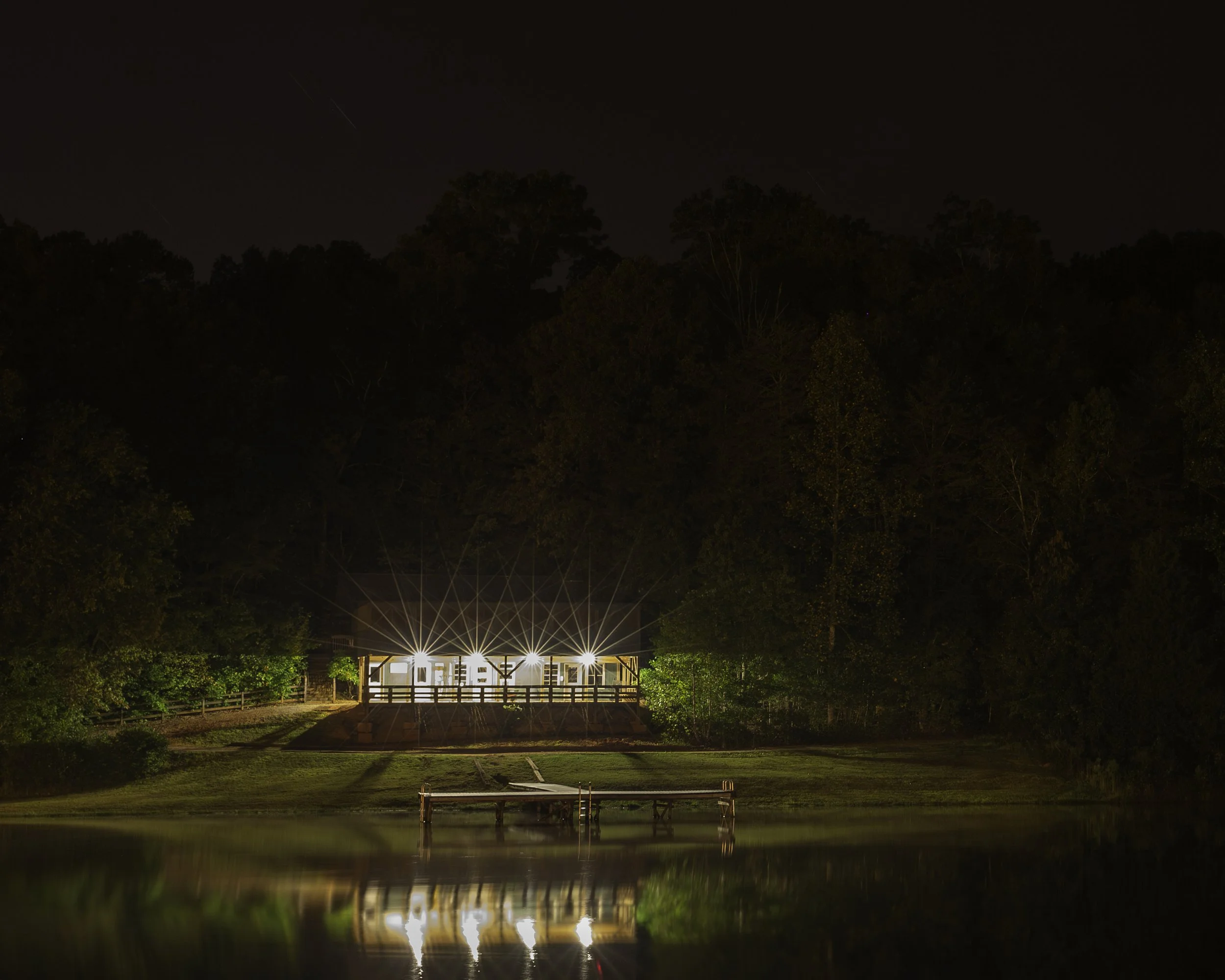 Nighttime view of a well-lit house on a hill surrounded by trees, with a dock extending into a calm body of water that reflects the house and lights.