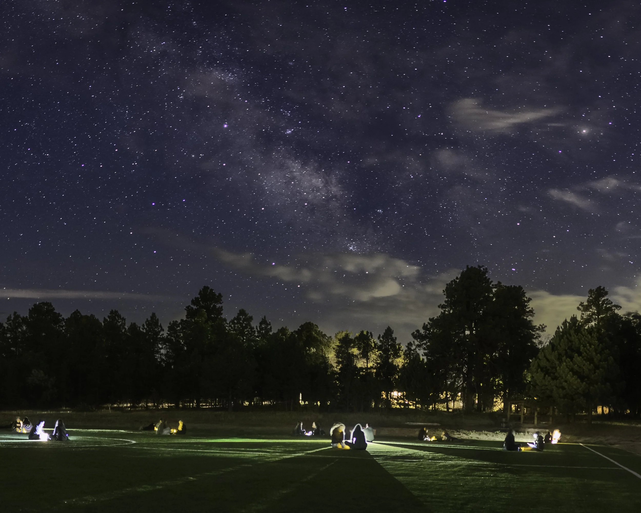 People sitting on the ground outdoors at night under a starry sky and visible the Milky Way galaxy, trees in the background, and some light illuminating the scene.