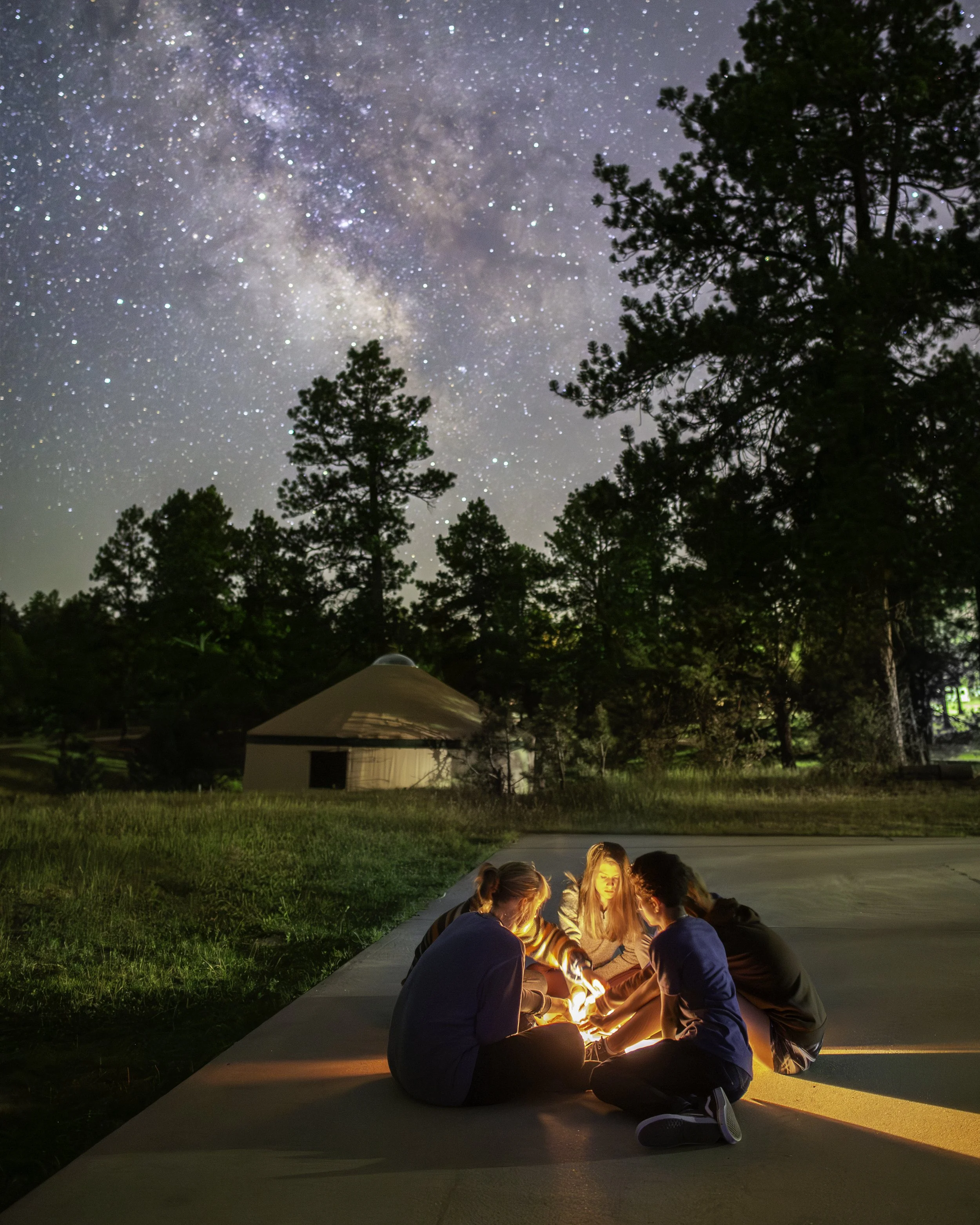 Group of children sitting on concrete pavement around a small campfire at night, under a starry sky with visible Milky Way, with trees and a dome-shaped building in the background.