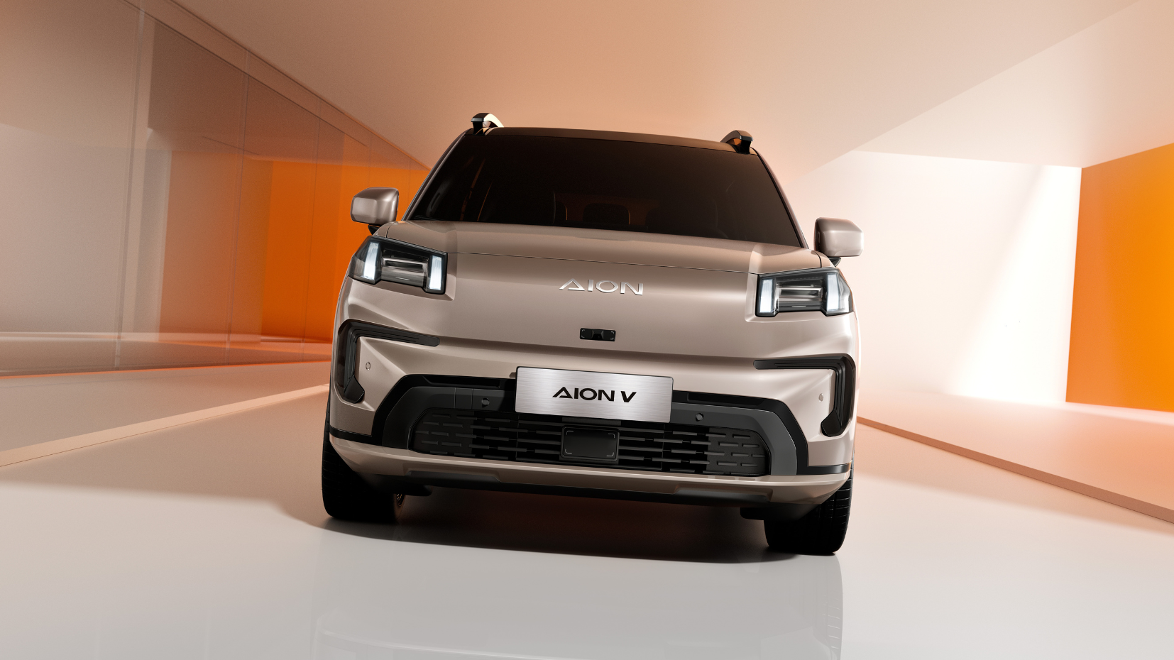 Front view of a modern, silver Aion V electric SUV in a minimalistic showroom with orange and white walls.