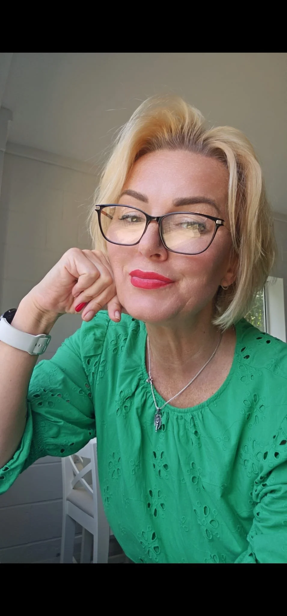 A middle-aged woman with blonde hair, wearing glasses, red lipstick, and a green embroidered blouse, sitting indoors with her chin resting on her hand.