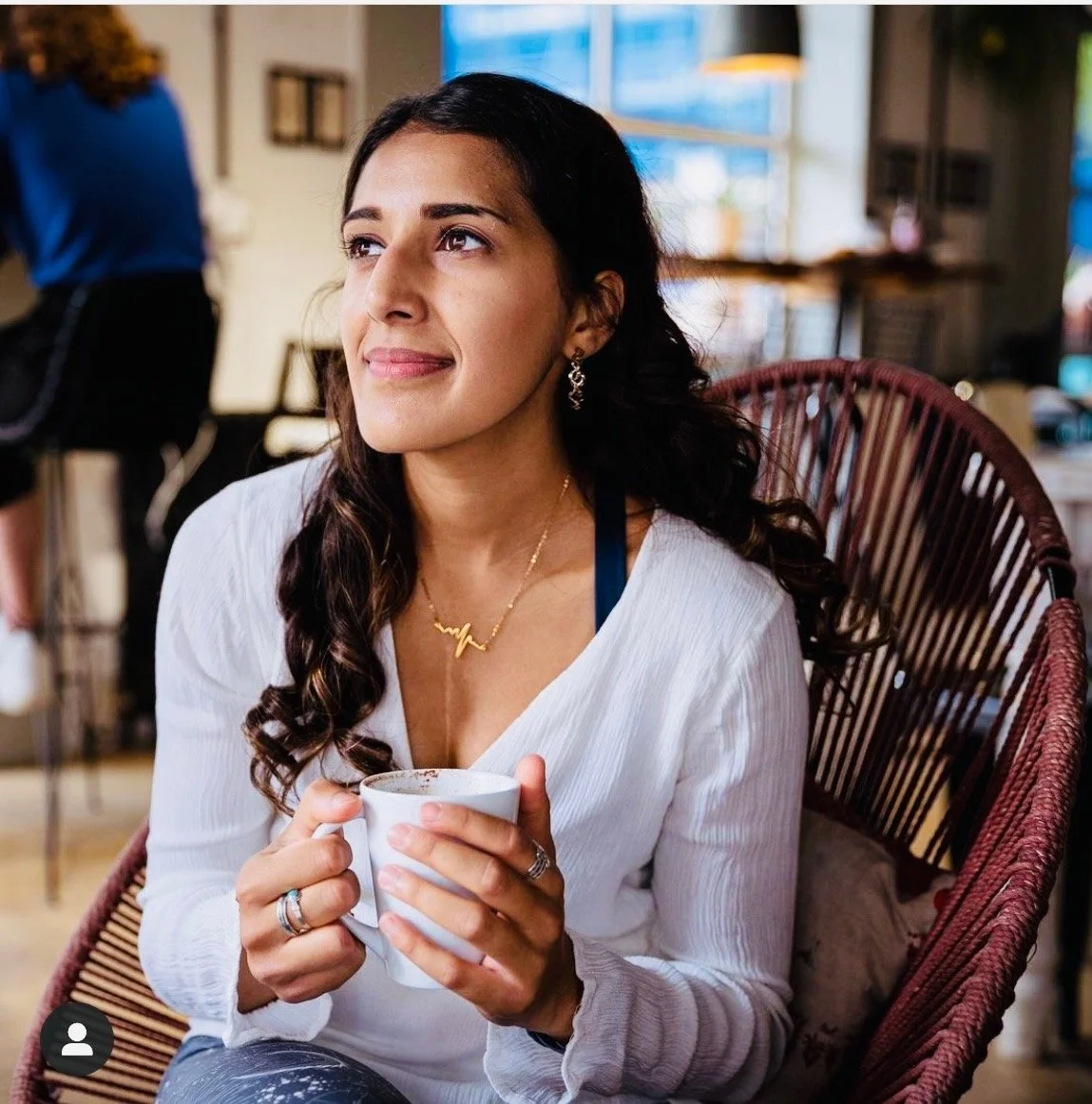 A woman with long curly dark hair and earrings is sitting in a cafe, holding a white mug, looking thoughtful. She is wearing a white blouse and a gold necklace with a heartbeat design.