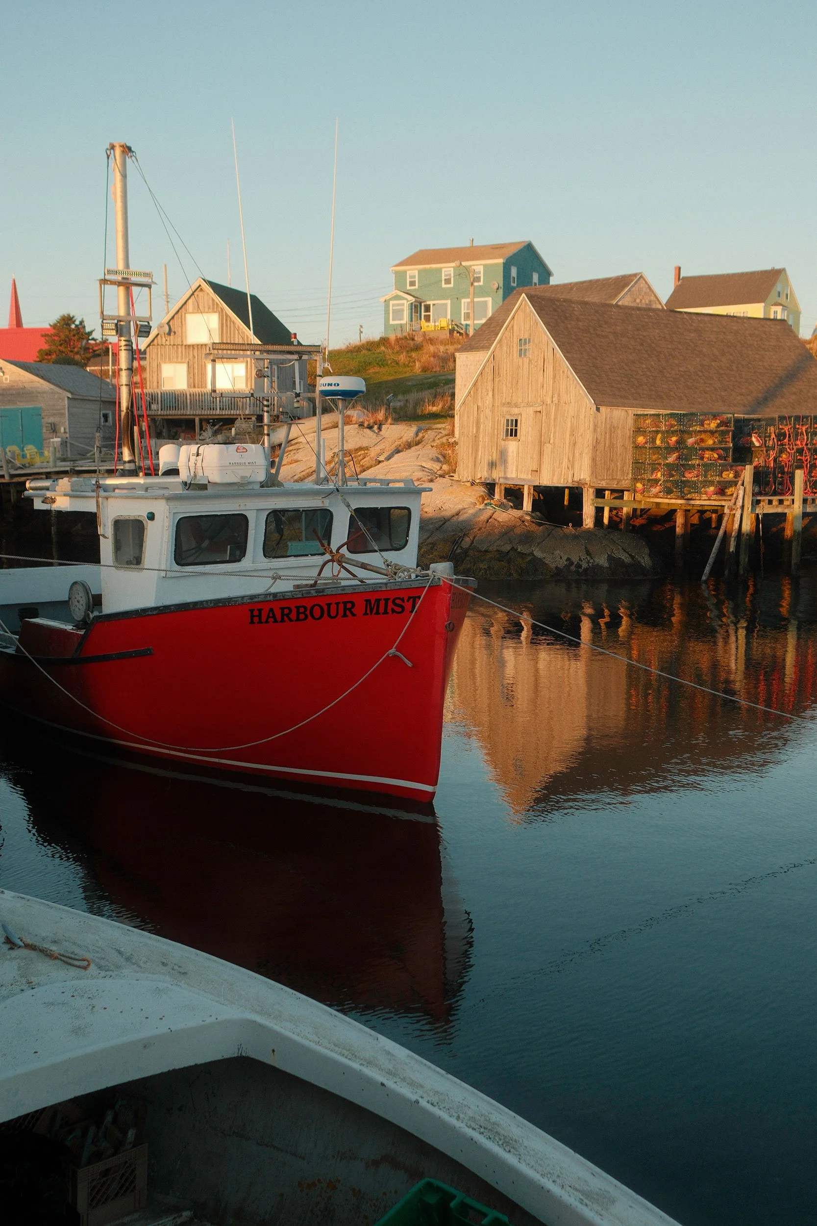 A red fishing boat named Harbour Mist is moored in Peggy's Cove with wooden fishing sheds, lobster traps, and colourful seaside homes in the background.