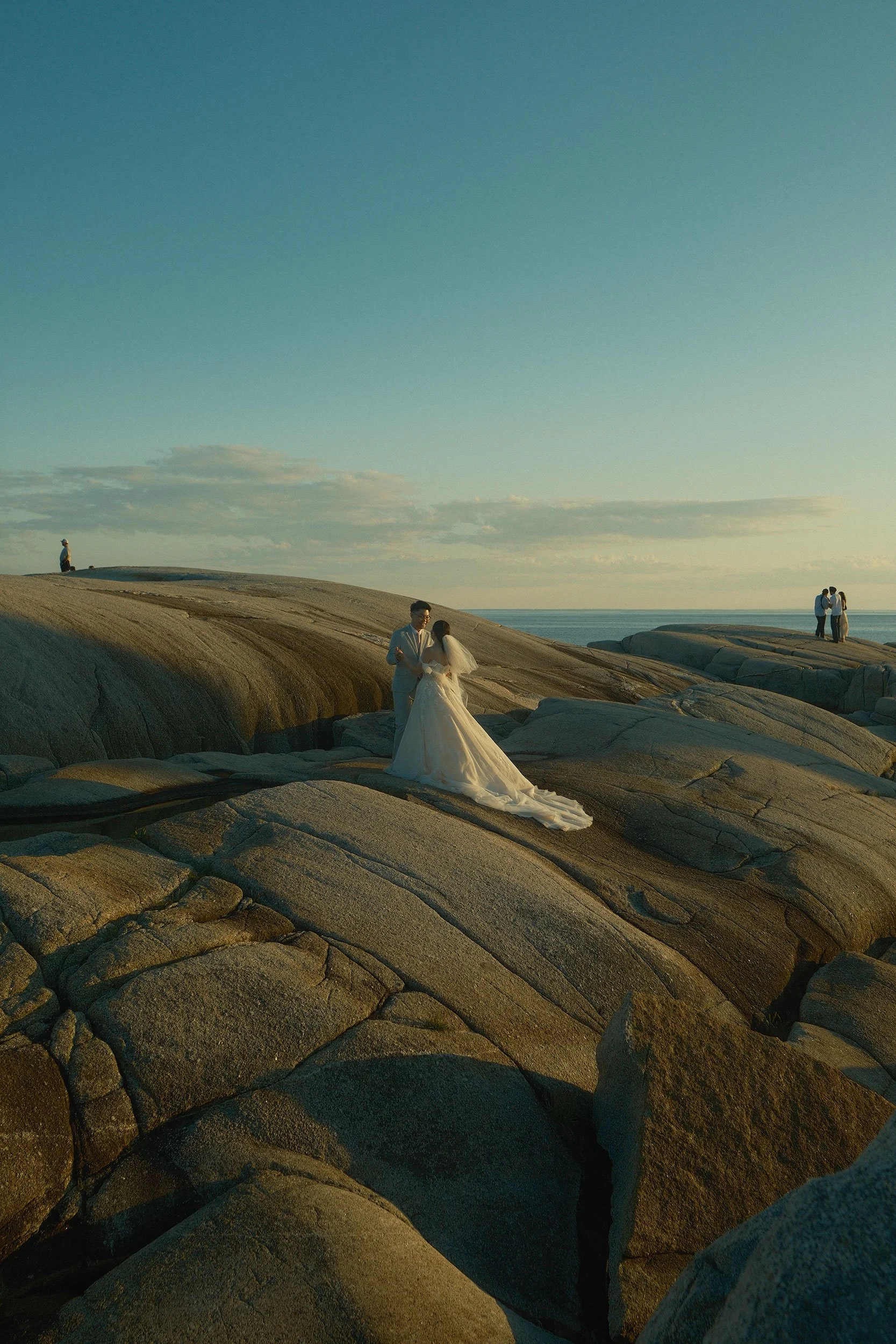 A couple in wedding attire standing on rocks near the ocean at Peggy's Cove. Other people in the background, at sunset or late afternoon.