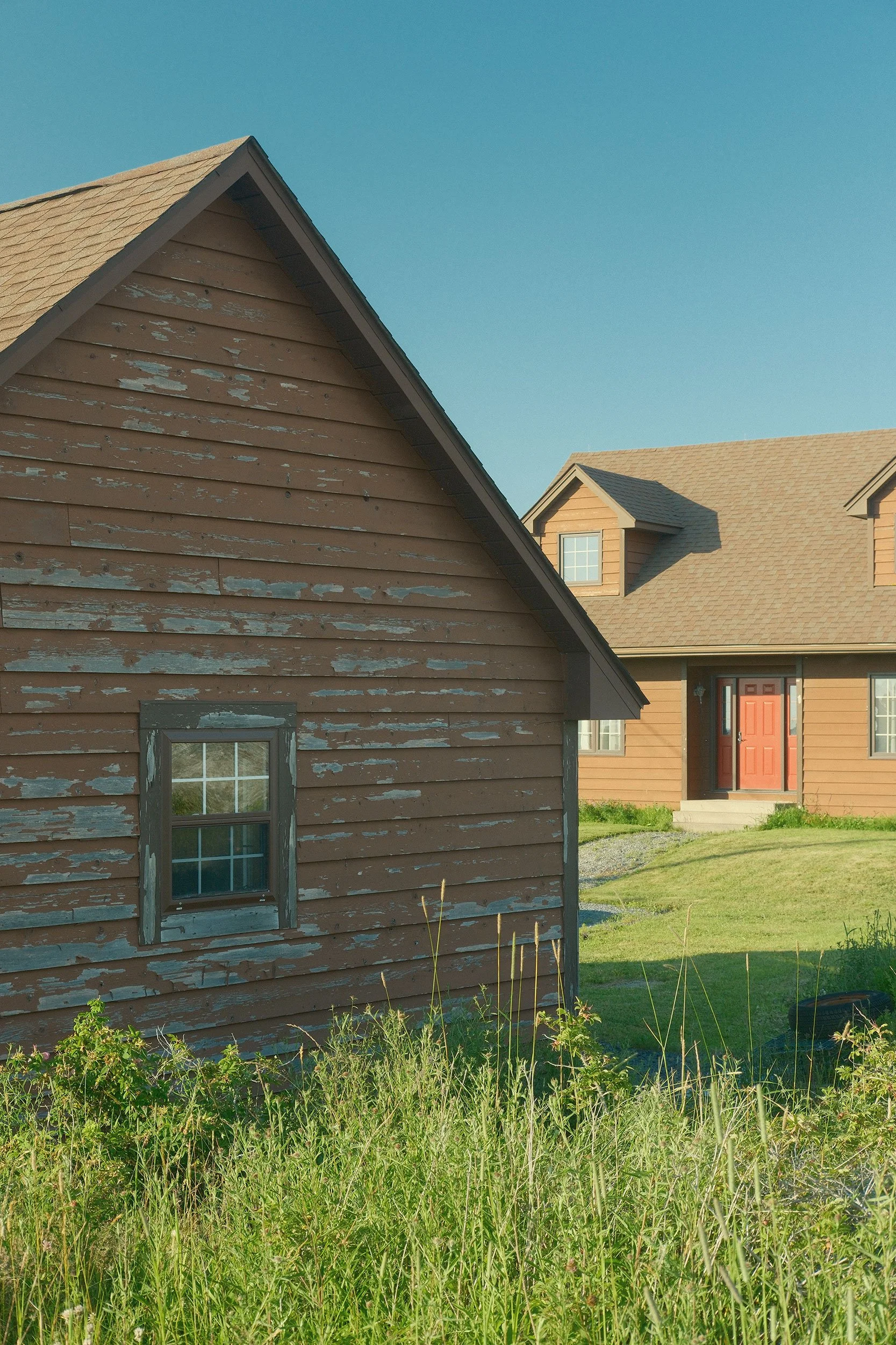 Front view of two houses on a sunny day, with green grass and plants in the foreground. The house in the foreground has weathered brown siding, while the background house has newer tan siding and orange door.