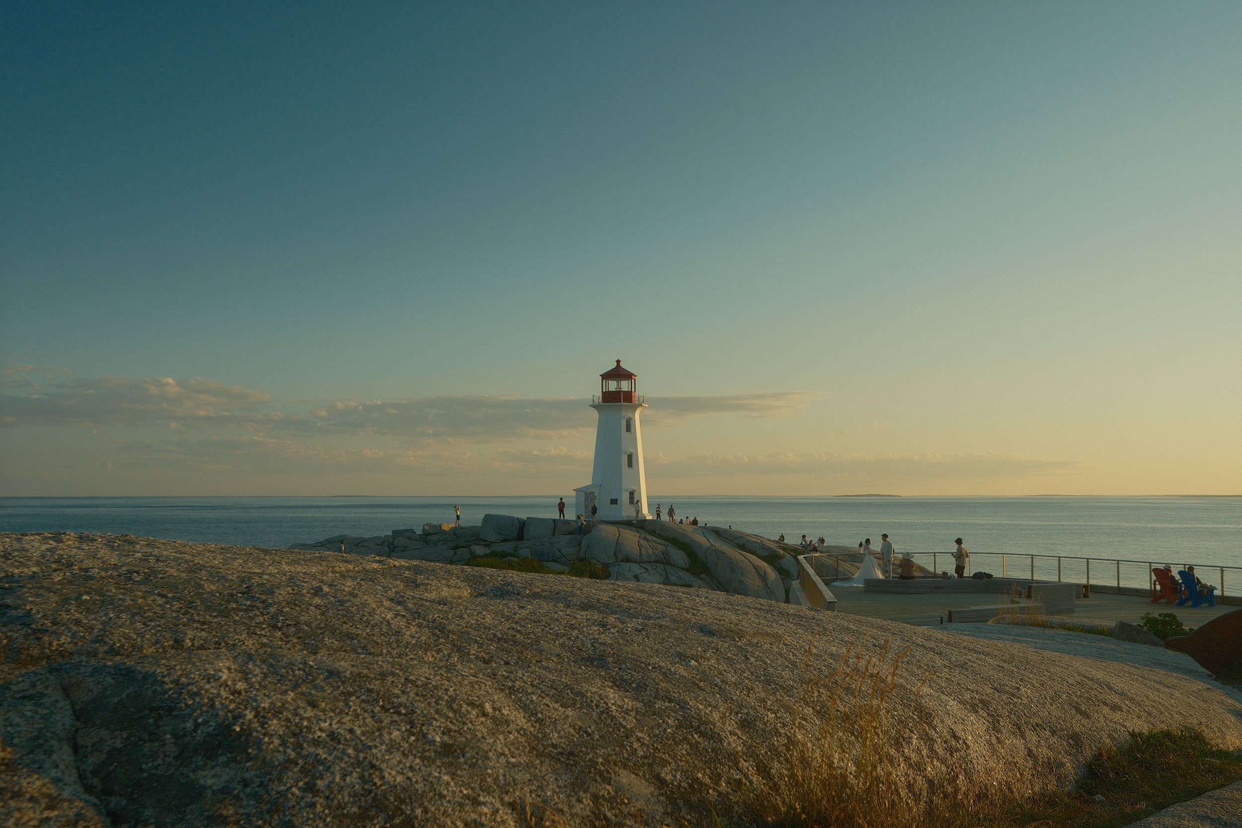 Peggy's Point lighthouse on the coast with a rocky foreground, calm water, and a clear sky with some clouds. People are walking and sitting near the lighthouse, enjoying the view.