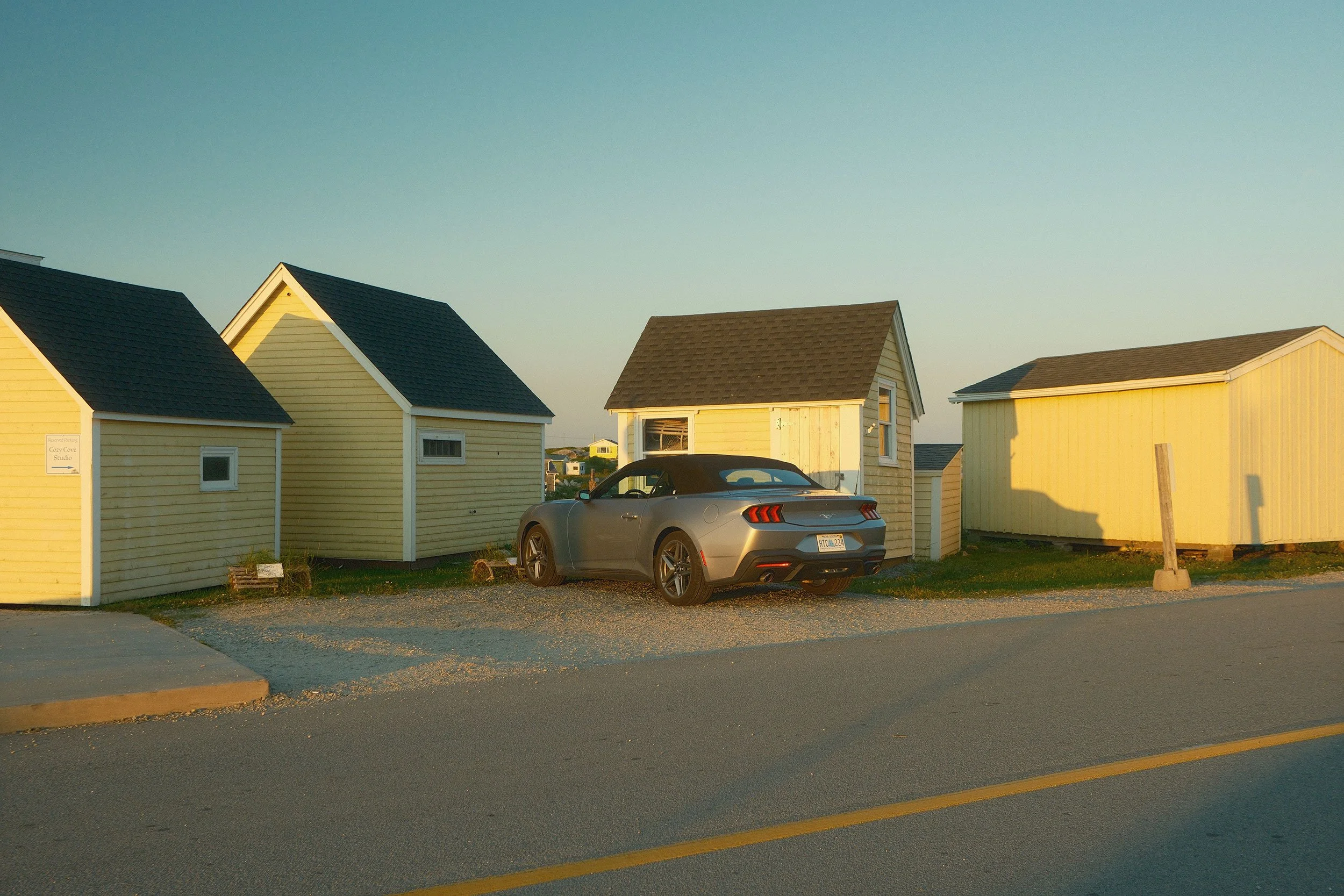 Small yellow shacks. A Ford Mustang convertible car parked in front, under a clear sky at sunset.