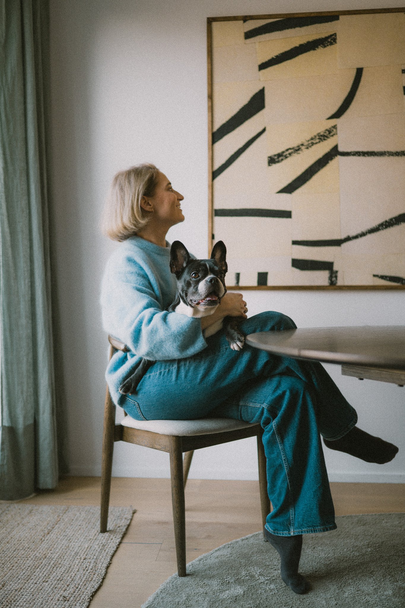 En kvinna sitter på en stol med en Bostonterrier hund i knät, tittar till höger och ler. Rummet har ett abstrakt konstverk på väggen och en gardin till vänster.