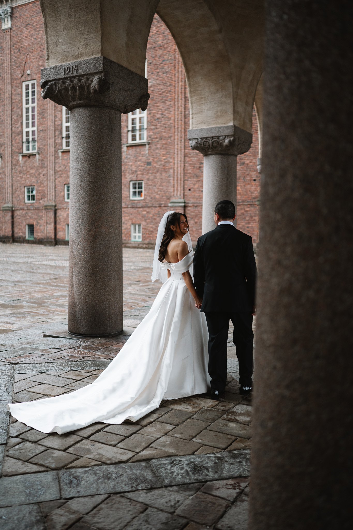 vigsel i stadshuset. En brud och brudgum håller handen och ser varandra i ögonen under en arkad, med en mörkbrun tegelbyggnad i bakgrunden.