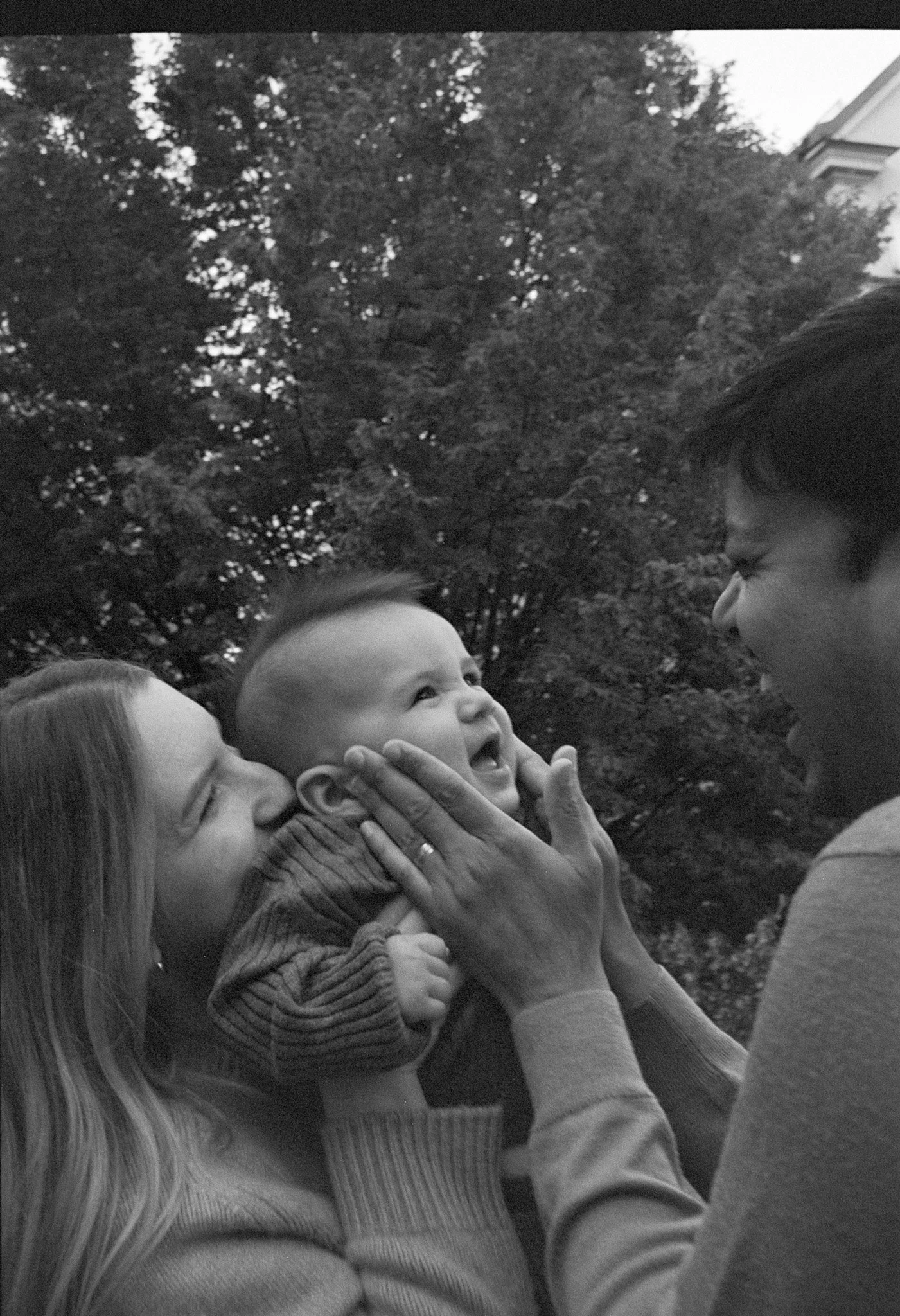 En kvinna håller en leende bebis medan en man trycker ansiktet mot bebisen i en utomhusmiljö med träd i bakgrunden.