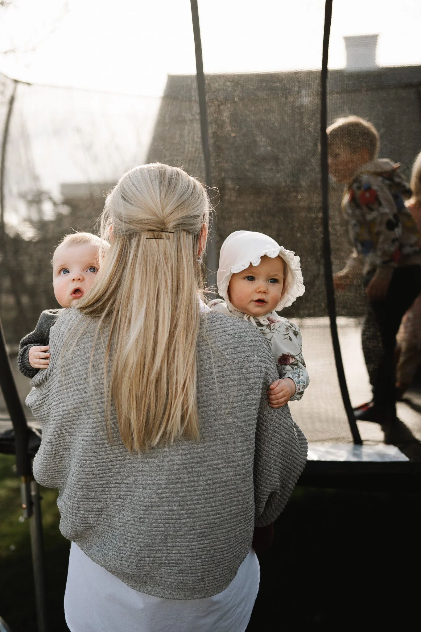 En kvinna håller två små barn, ett i varje arm, utomhus framför ett skyddsnät. Barnen tittar nyfikt.