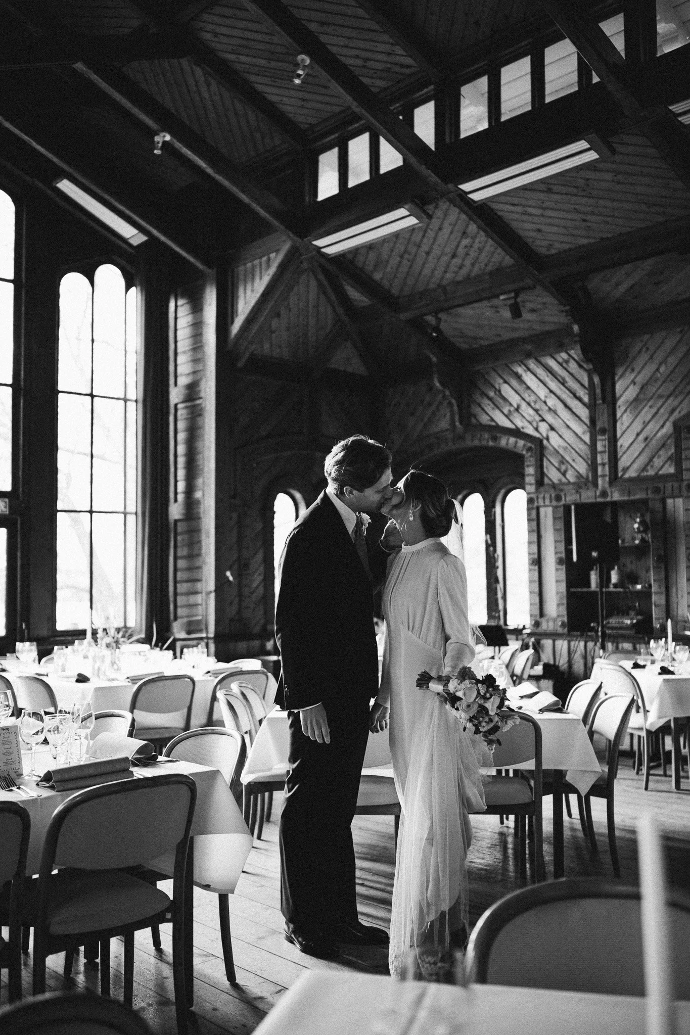 carlshälls gård i stockholm.Ett monochromt foto av ett bröllopsceremoni med ett par som kysser varandra i ett rustikt inomhusrum med träväggar och stora fönster.
