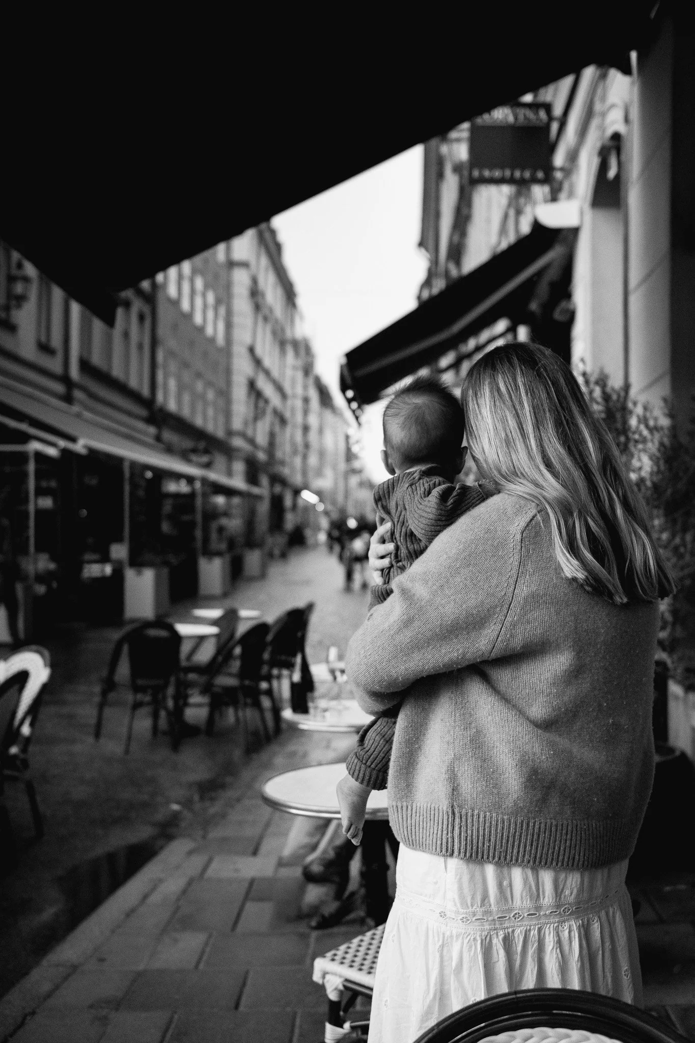 En kvinna håller ett barn i en stadsgata med uteservering och tomma stolar, svartvitt.