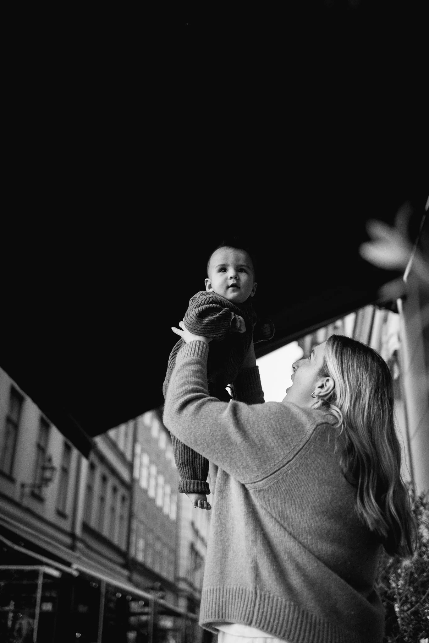 En kvinna lyfter en bebis i luften på en stadsamhörning, svartvitt foto.