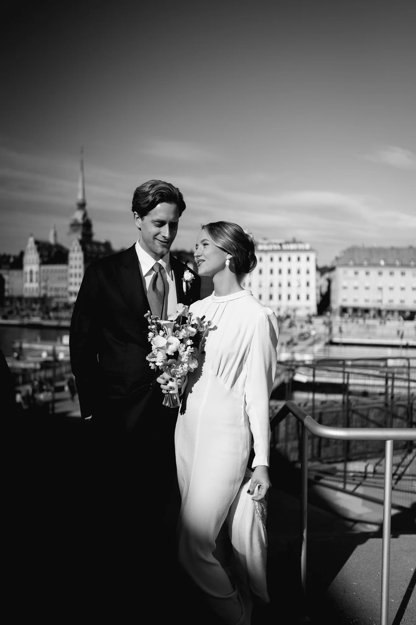 södermalm bröllop.Svartvitt foto av ett brudpar utomhus, med stadsvyer i bakgrunden, på en klar dag.