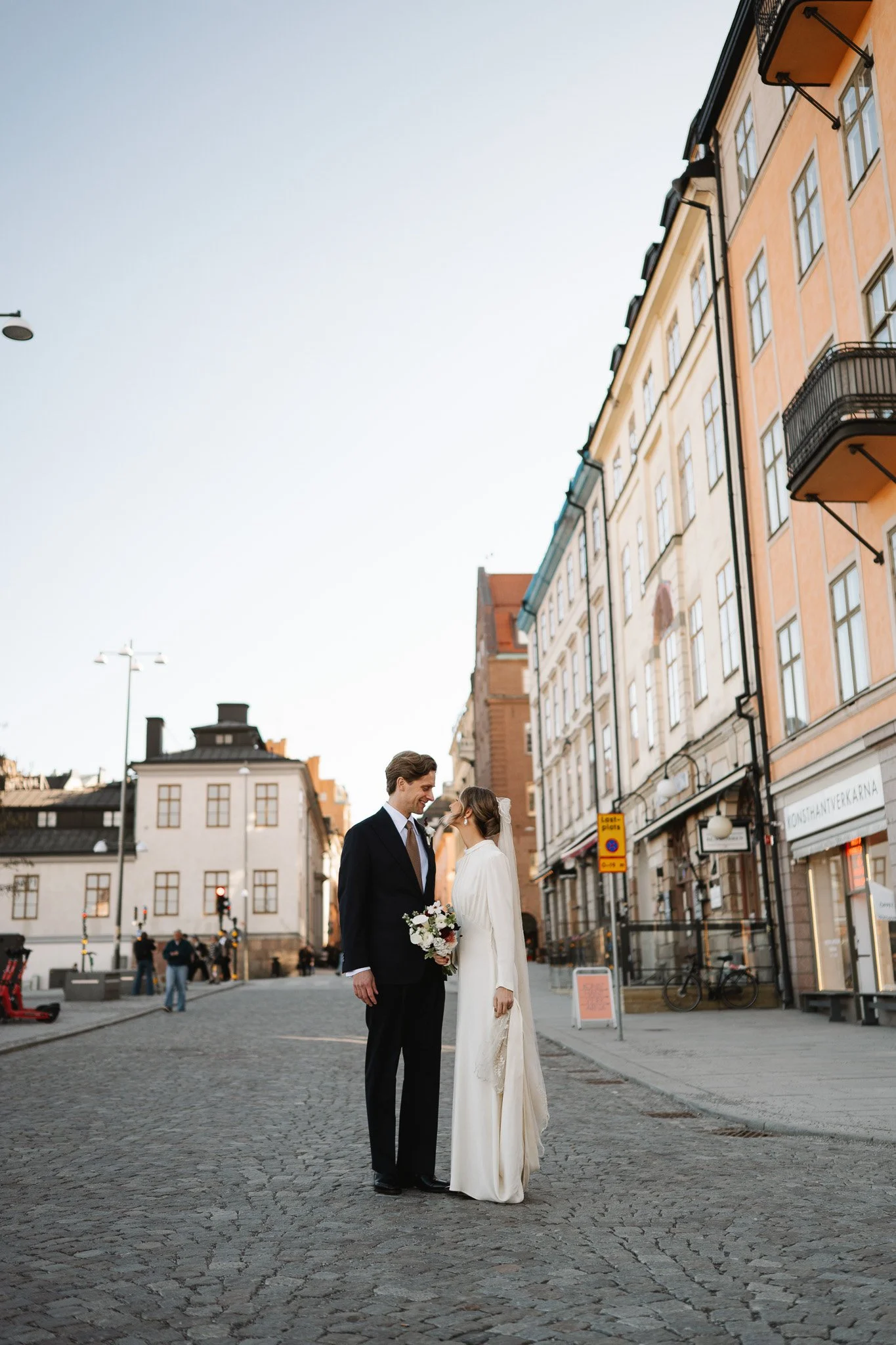 södermalm bröllop.En nygift brud och brudgum som står på en gatukorsning i en stad, med gamla byggnader i bakgrunden, under en klar himmel.
