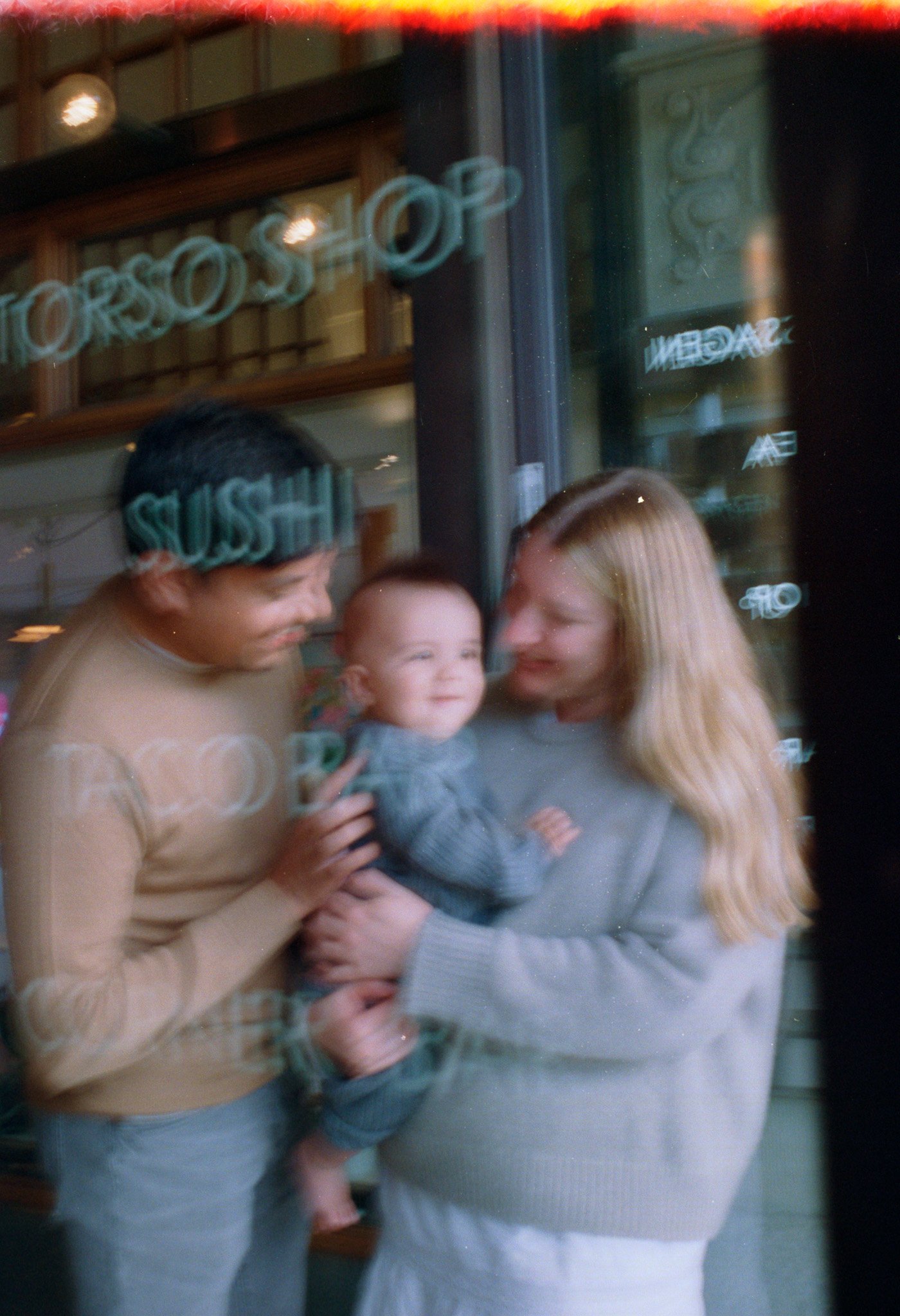 Två kvinnor som håller en leende bebis inomhus, synlig genom ett fönster med texten 'SUSHI'.