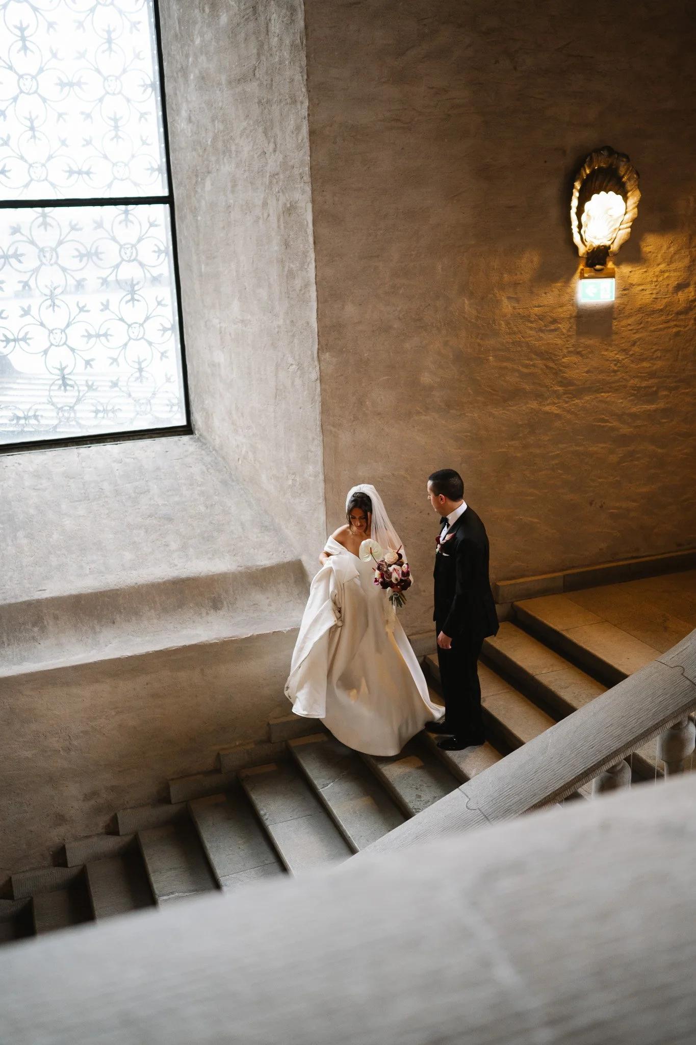 vigsel i stadshuset fotografi.En brud och en brudgum står på en trappa inomhus, bruden bär en vit brudklänning och håller en bukett medan brudgummen bär en svart kostym.