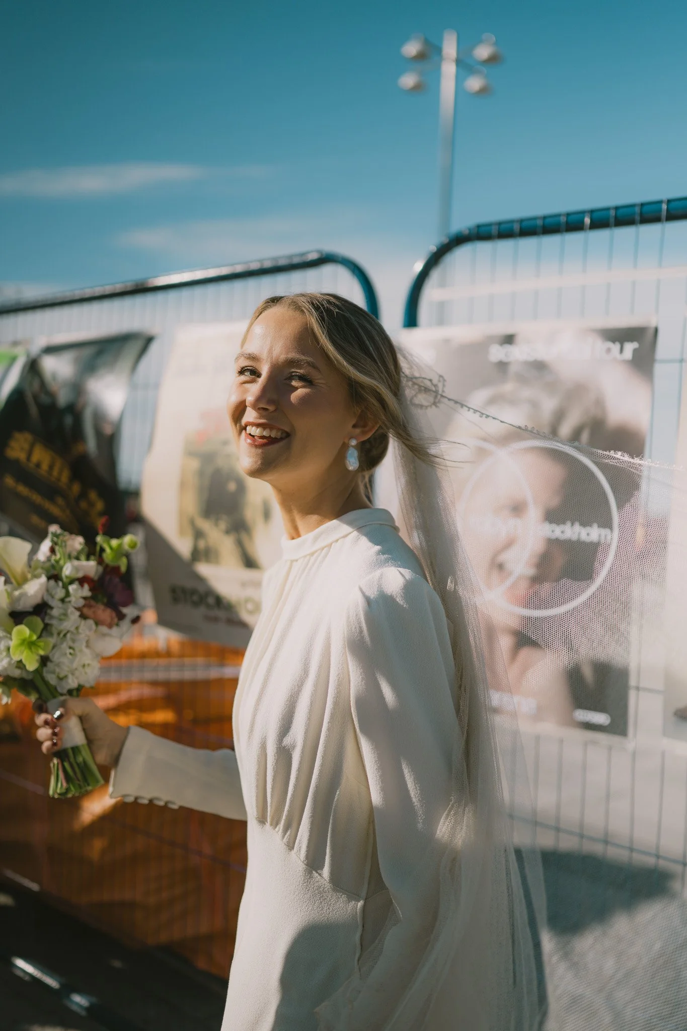 En glad kvinna i en vit klänning med en blomsterbukett står utomhus i solsken, med en metallstaket som bakgrund. södermalm bröllop.