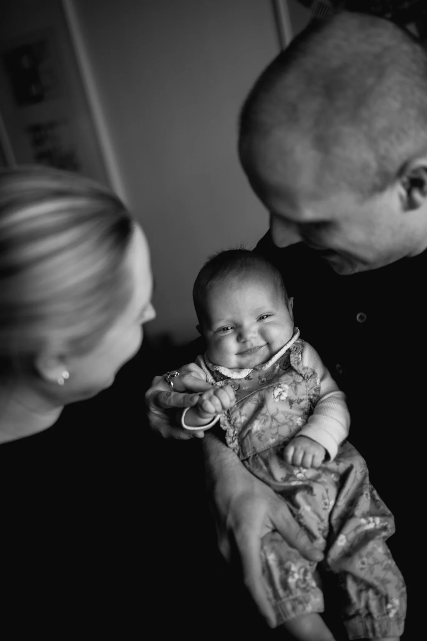 En leende baby som hålls av en man, medan en kvinna tittar på dem.