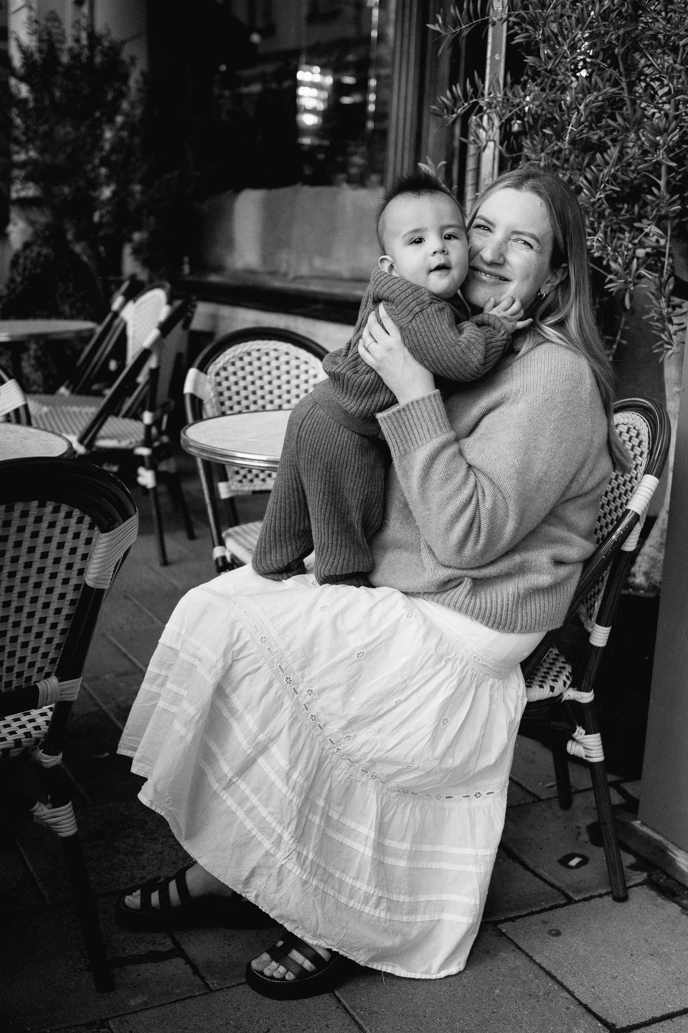 En kvinna i en kjol och tröja som sitter på en stol på en uteservering, håller ett barn i en tröja och byxor, ler och myser tillsammans.