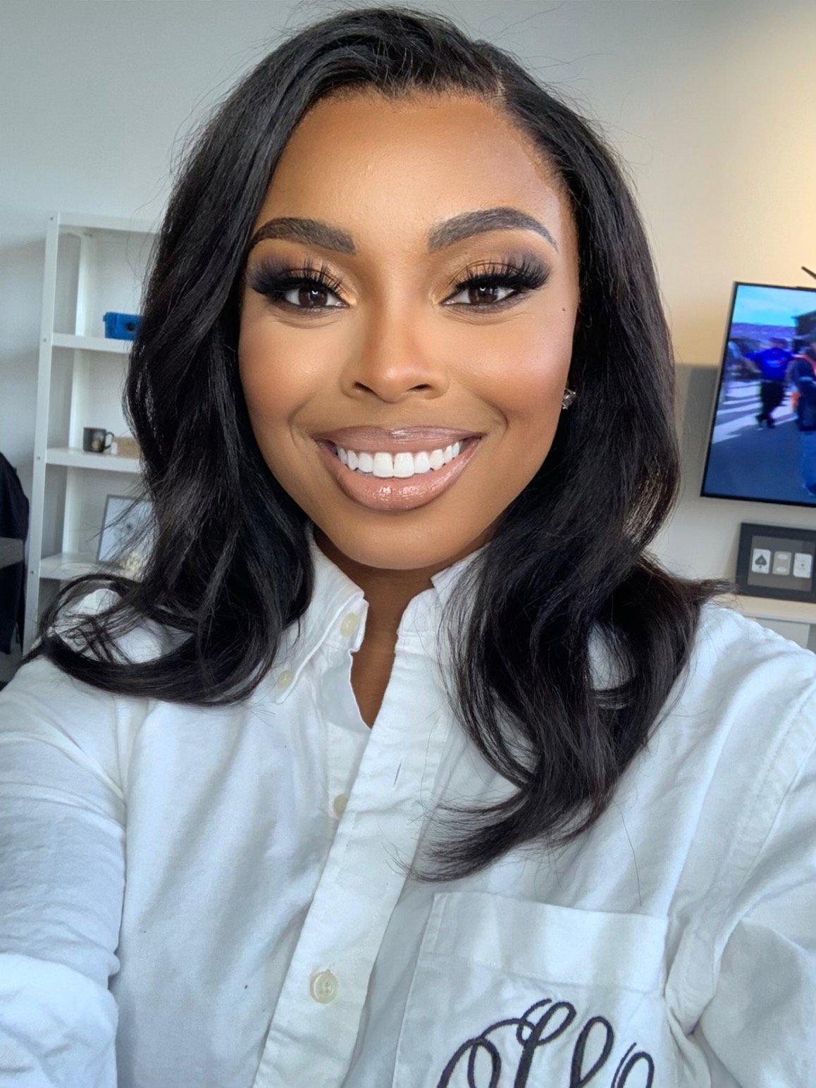 Smiling woman wearing a white shirt, with a bookshelf and TV in the background.