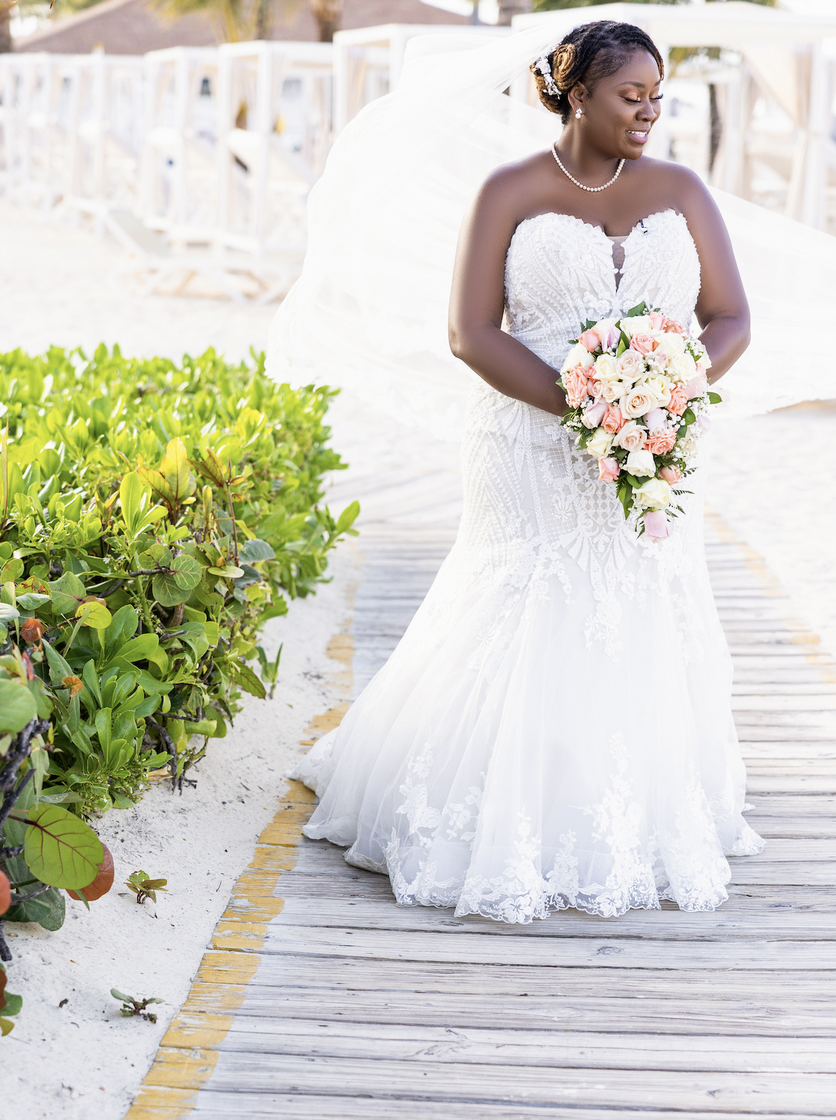 Bride in white lace wedding dress holding a bouquet on a wooden boardwalk at the beach.