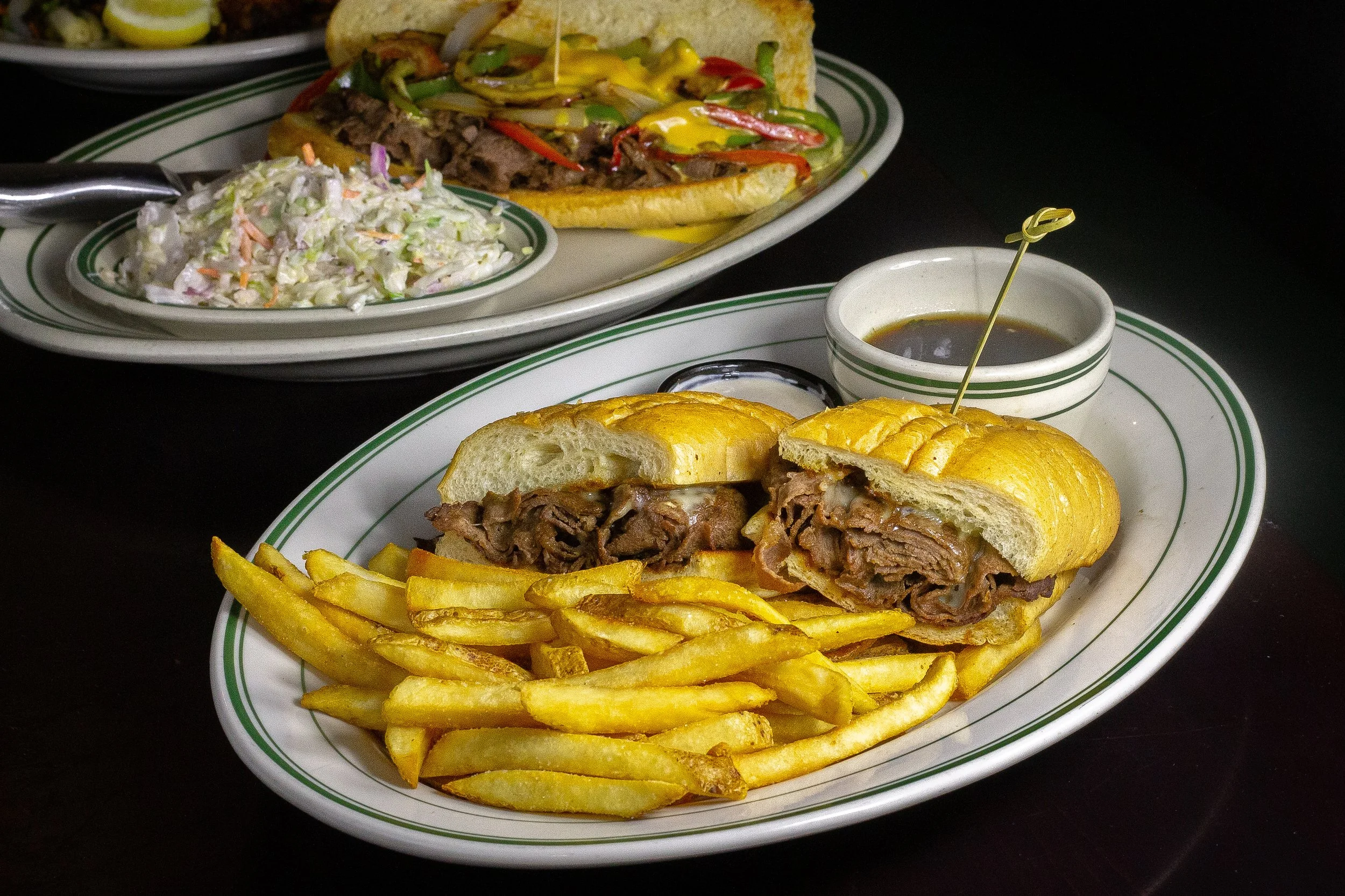 A plate with a beef and french fry sandwich, a side of French fries, and a small bowl of gravy, with another dish of salad and a specialty hot dog in the background.