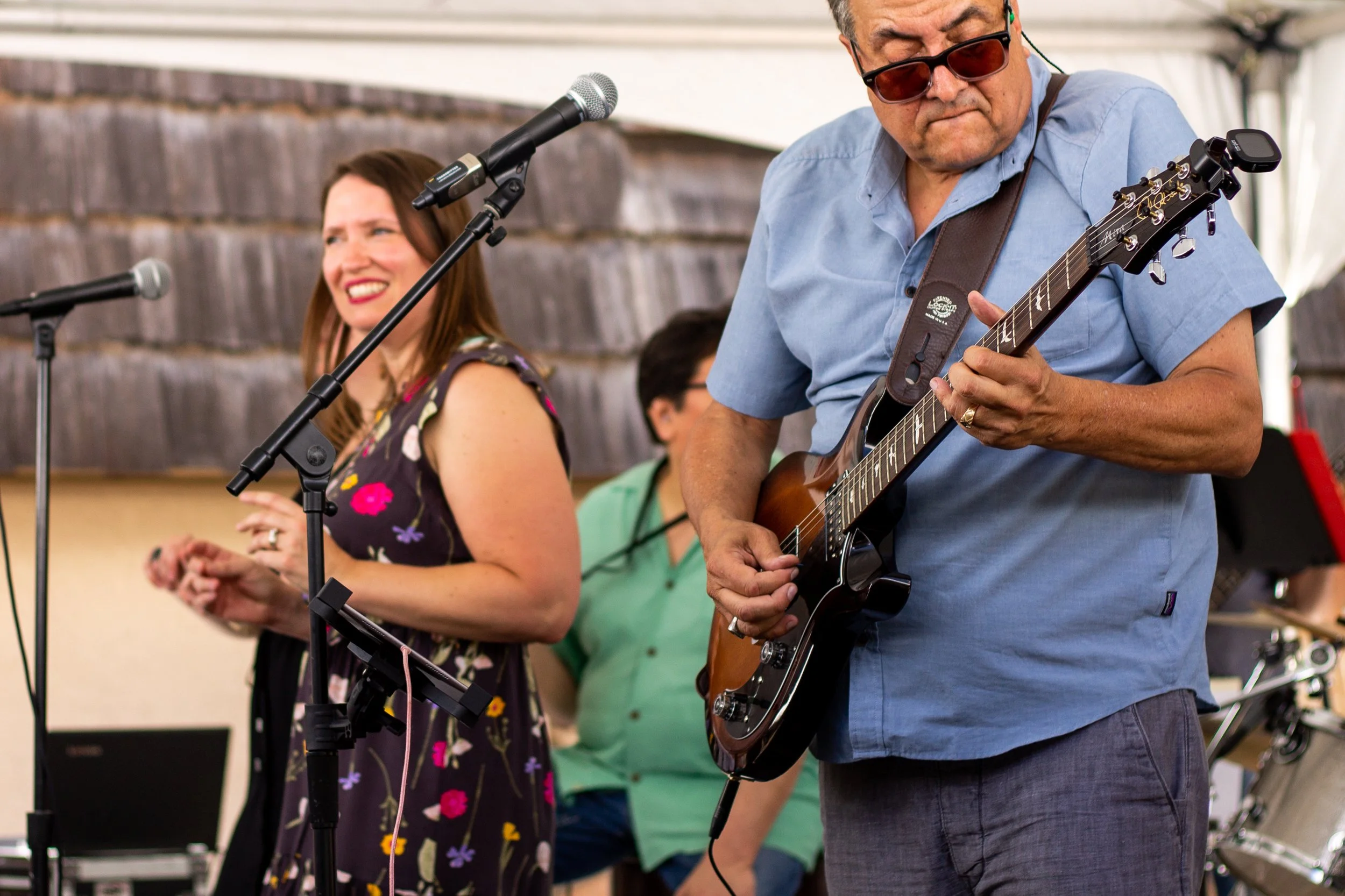 Musicians performing outdoors, featuring a woman singing into a microphone and a man playing an acoustic guitar, with people in the background.