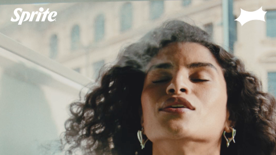 Mulher com cabelo cacheado, fechando os olhos, na rua com edifícios ao fundo, com logo da Sprite na parte superior esquerda.