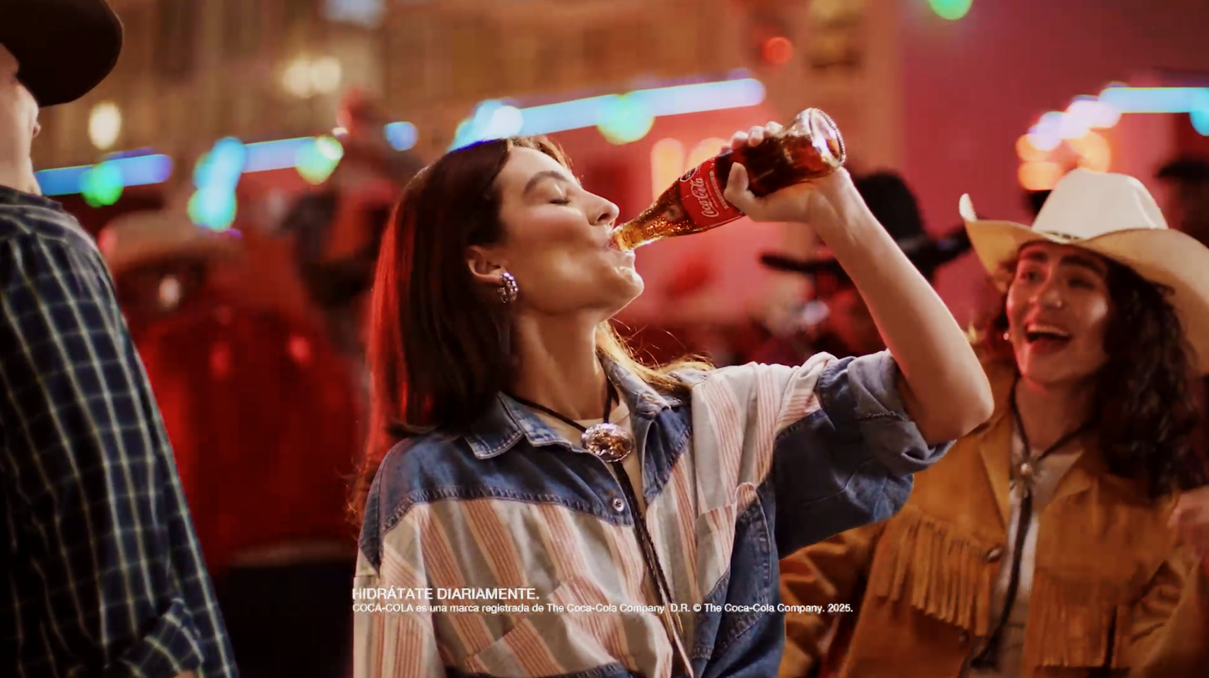 Pessoas em uma festa com luzes coloridas, uma mulher bebe refrigerante Coca-Cola de uma garrafa, usando roupa jeans listrada, e outra mulher com chapéu de cowboy e roupa de couro. Ambos estão sorrindo.