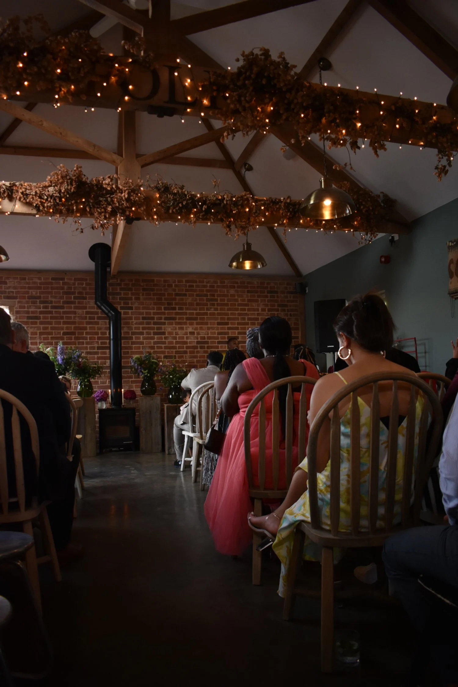 Indoor wedding ceremony with guests seated, rustic decor with dried flowers and string lights, brick wall backdrop, wooden chairs.