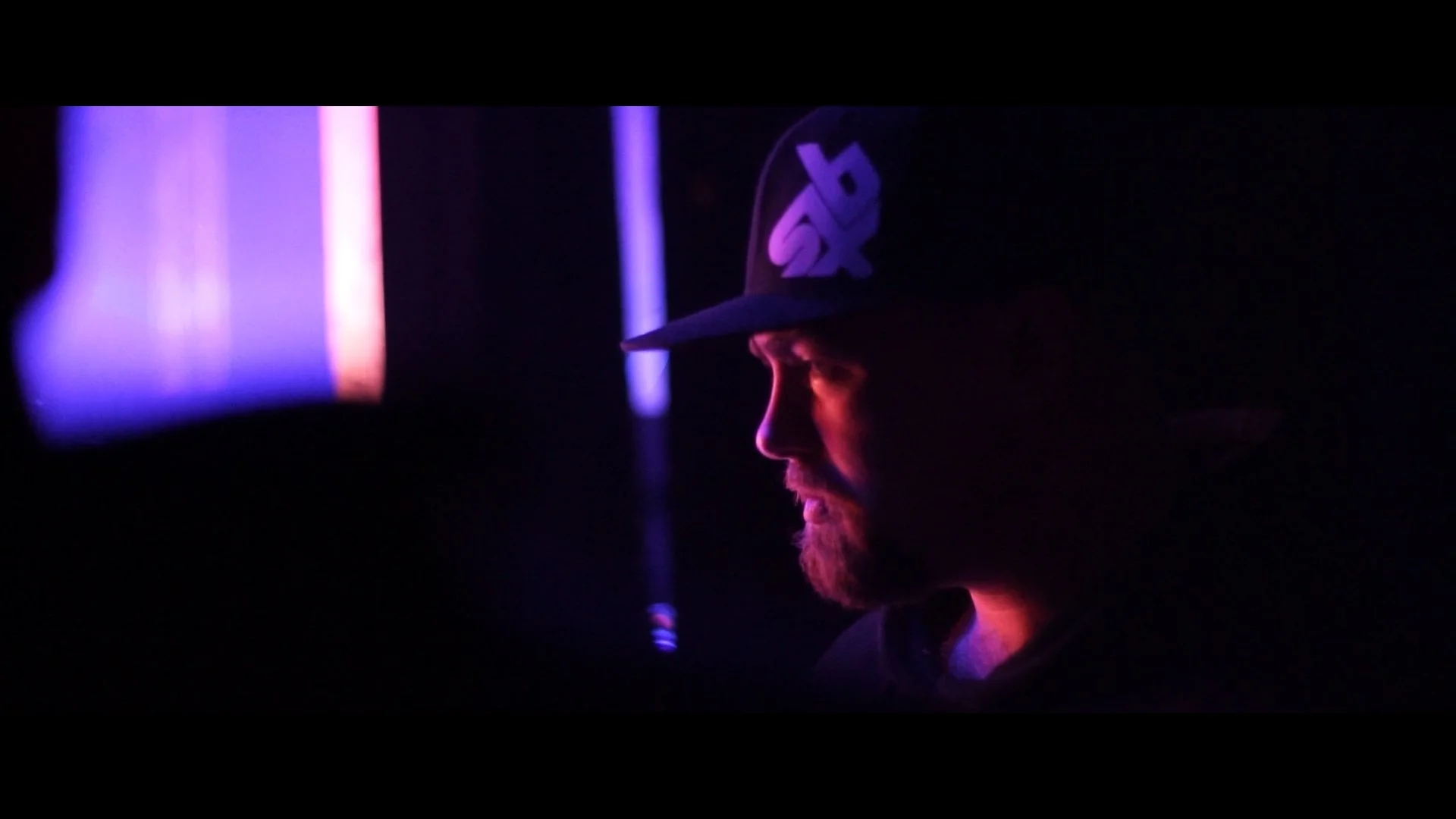 Nahaufnahme eines Mannes mit Bart und Baseballkappe in dunkler Umgebung, beleuchtet von bunten Neonlichtern.