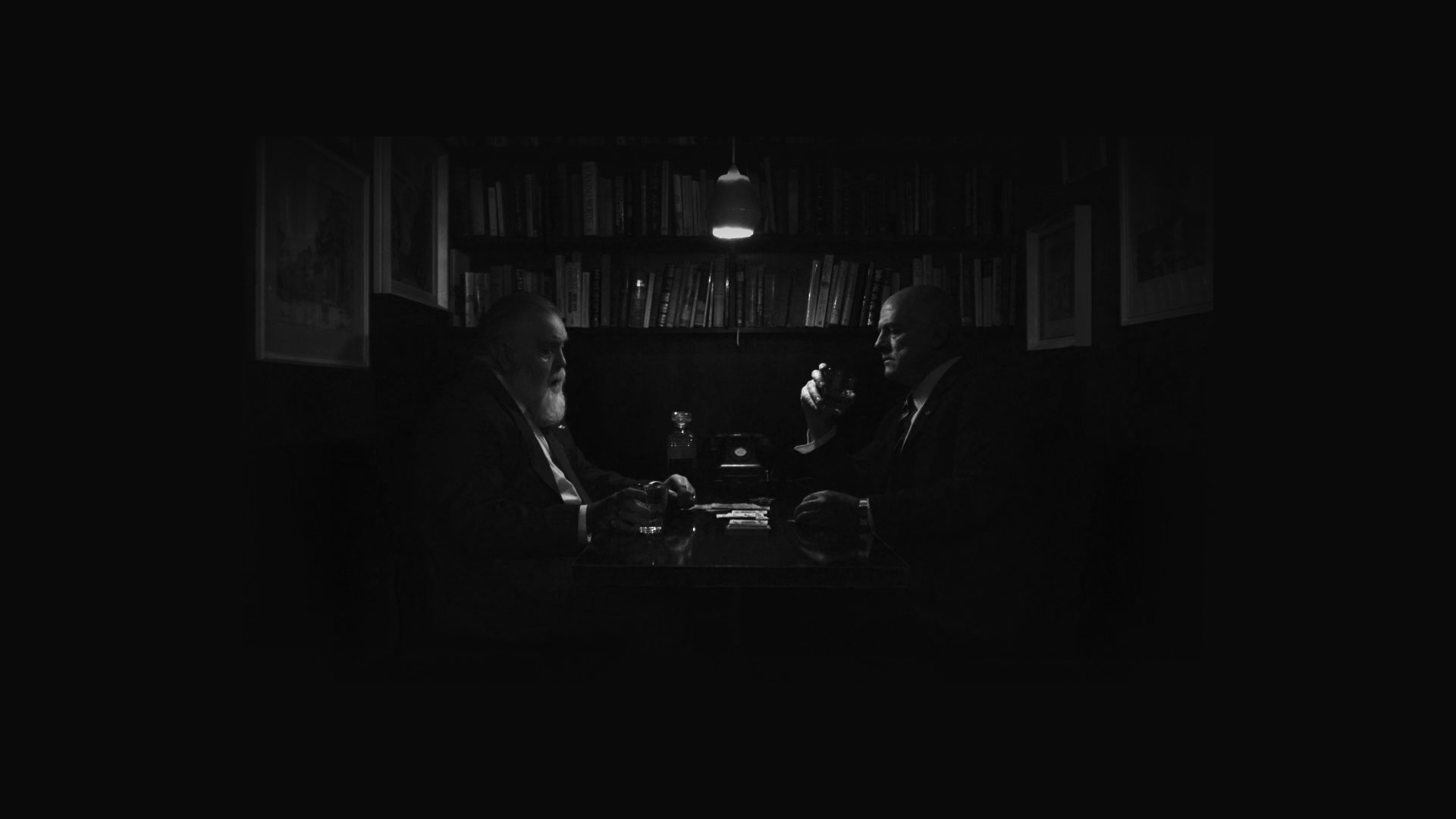 Zwei Männer in Anzügen sitzen sich in einem dunklen Raum an einem Tisch gegenüber, einer trinkt aus einem Glas, im Hintergrund Bücherregale und eine Lampe.