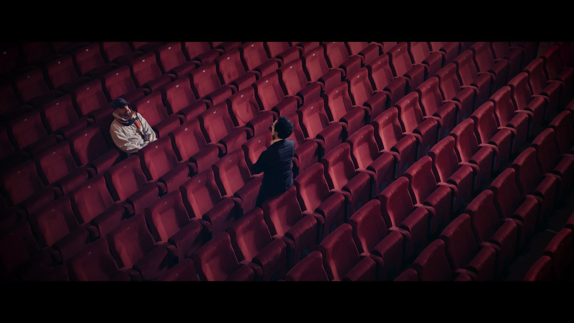Zwei Personen sitzen mit Abstand in einem leeren Theater mit roten Plätzen.