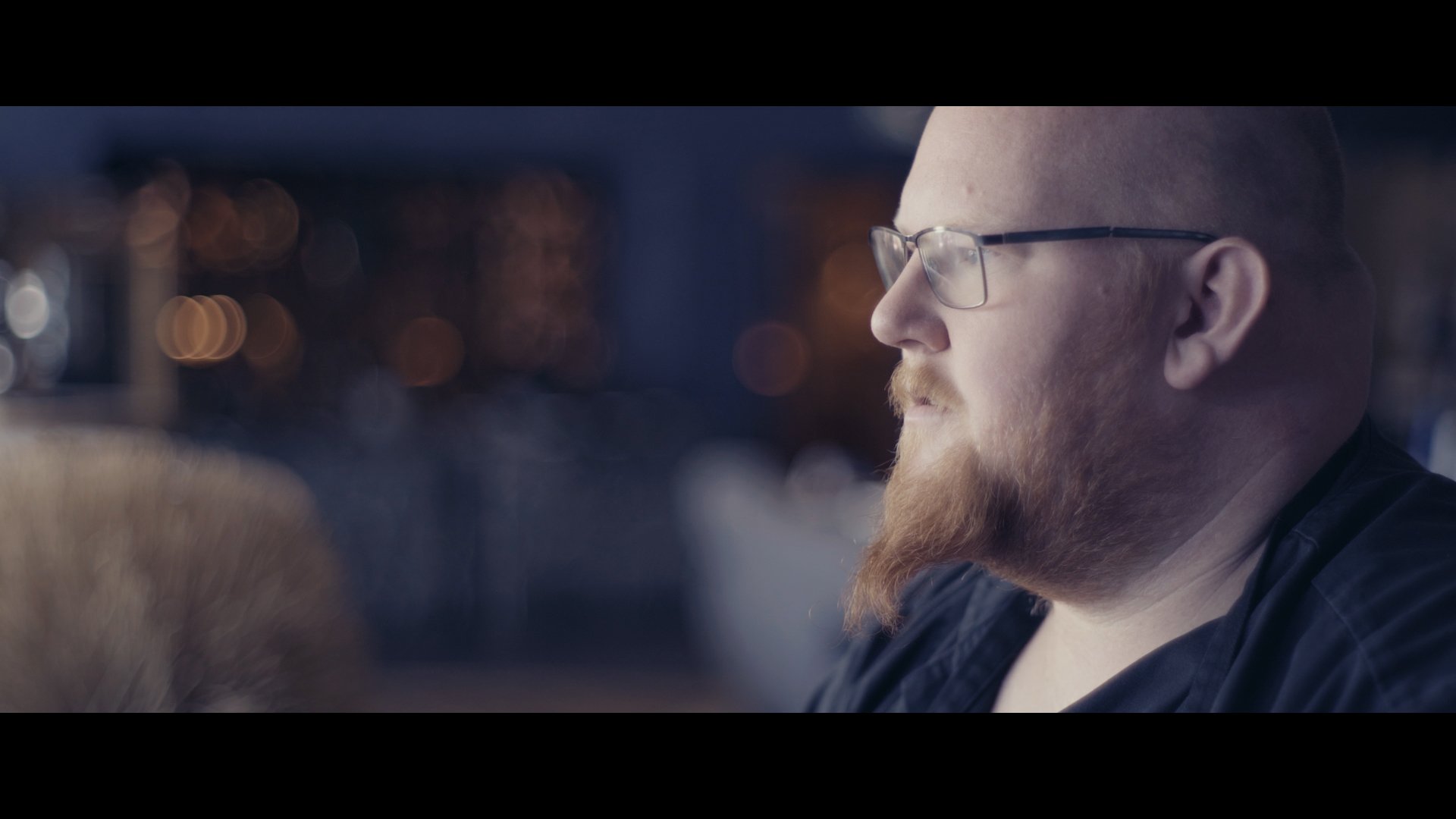 Nahaufnahme eines Mannes mit Brille, der nach rechts blickt, im Hintergrund unscharfe Lichter bei Nacht.