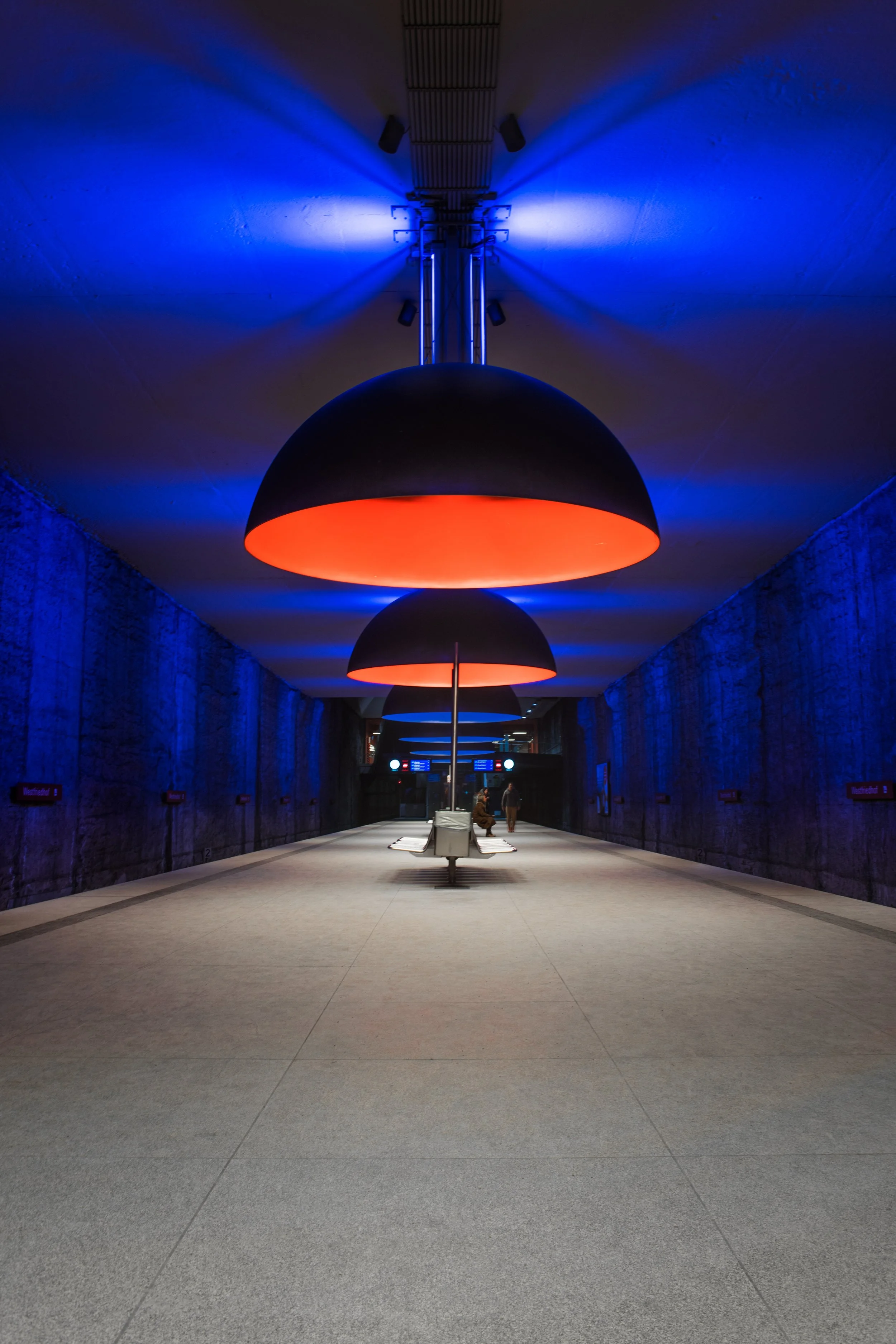 Das Bild zeigt einen unterirdischen Bahnhof mit blauen und roten Lichtinstallationen an der Decke, eine Sitzbank in der Mitte und einige wartende Menschen.