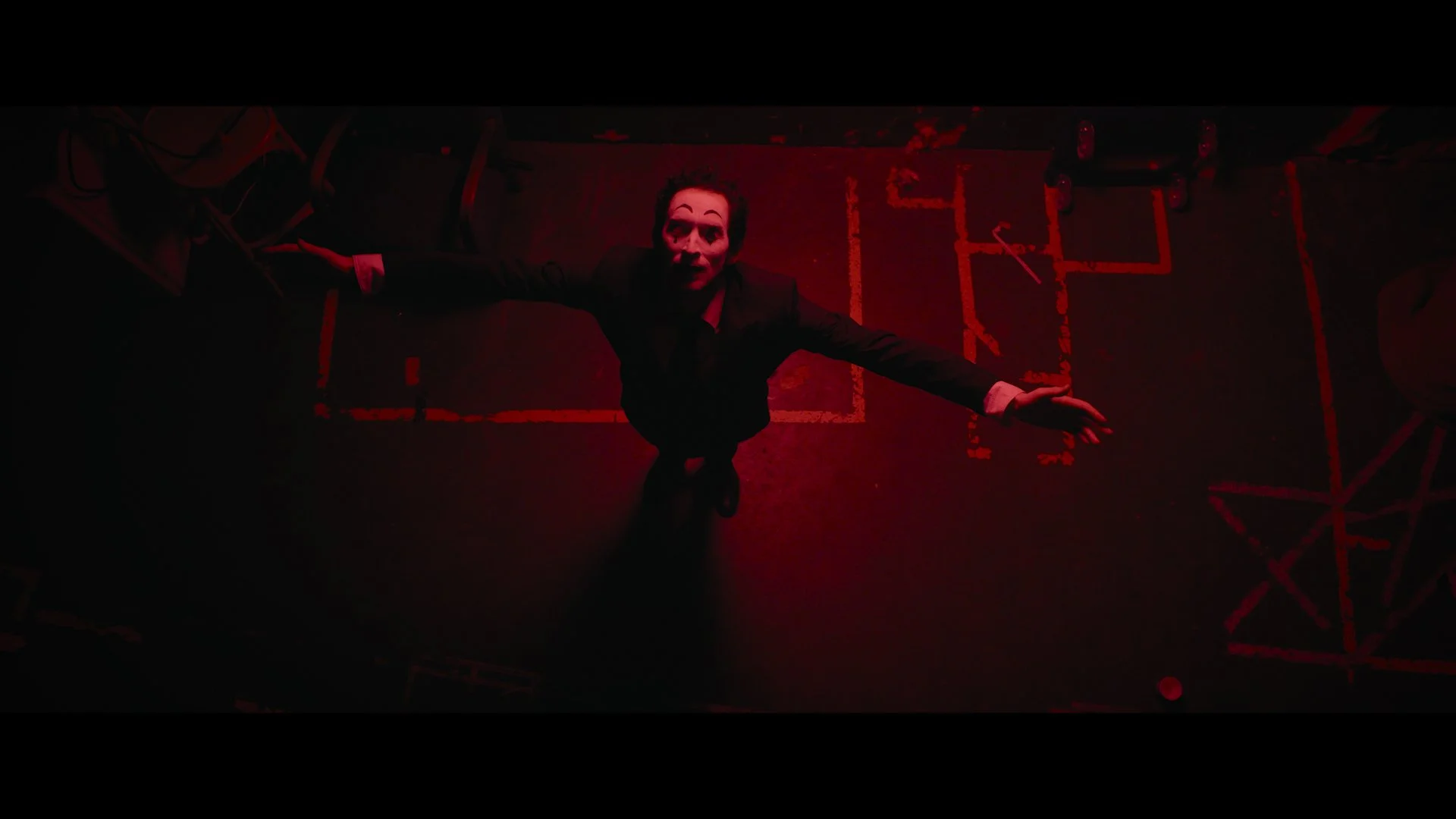 Eine Figur in einem schwarzen Anzug liegt auf dem Boden in einem dunklen Raum mit roten Lichtern, die auf den Boden gemalt sind, und kleinen Gegenständen auf der Seite.